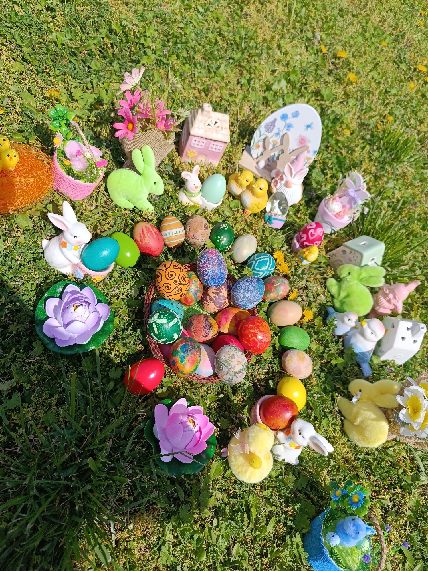 The image shows an Easter display arranged on a patch of grass in an outdoor setting. The display includes various Easter-themed decorations such as Easter eggs, ceramic and plush bunnies, small chicks, floral arrangements, and miniature decorative houses. The Easter eggs, many with colorful painted designs, are scattered around or placed in a woven basket. The grass appears to be green and well-maintained. The photo was likely taken in daylight due to the strong sunlight and shadows. The location is Cherkovitsa, Bulgaria.
