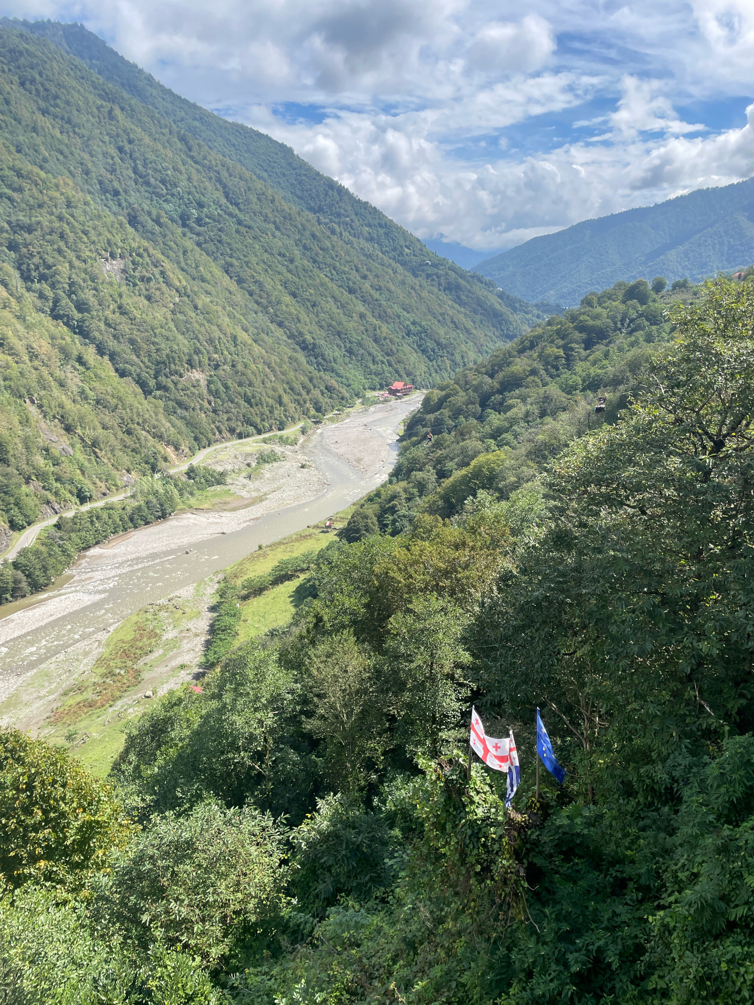 The image captures a wide shot of a verdant mountain valley in Gvara, Georgia. A river winds through the valley floor, flanked by a road that follows its course. Steep, tree-covered mountains rise on either side, with some clouds visible in the sky. In the foreground, two flags are prominently displayed on a hillside overlooking the valley: the flag of Georgia and the flag of the European Union. There are also cable cars visible in the distance, suggesting a tourist attraction or transportation system.