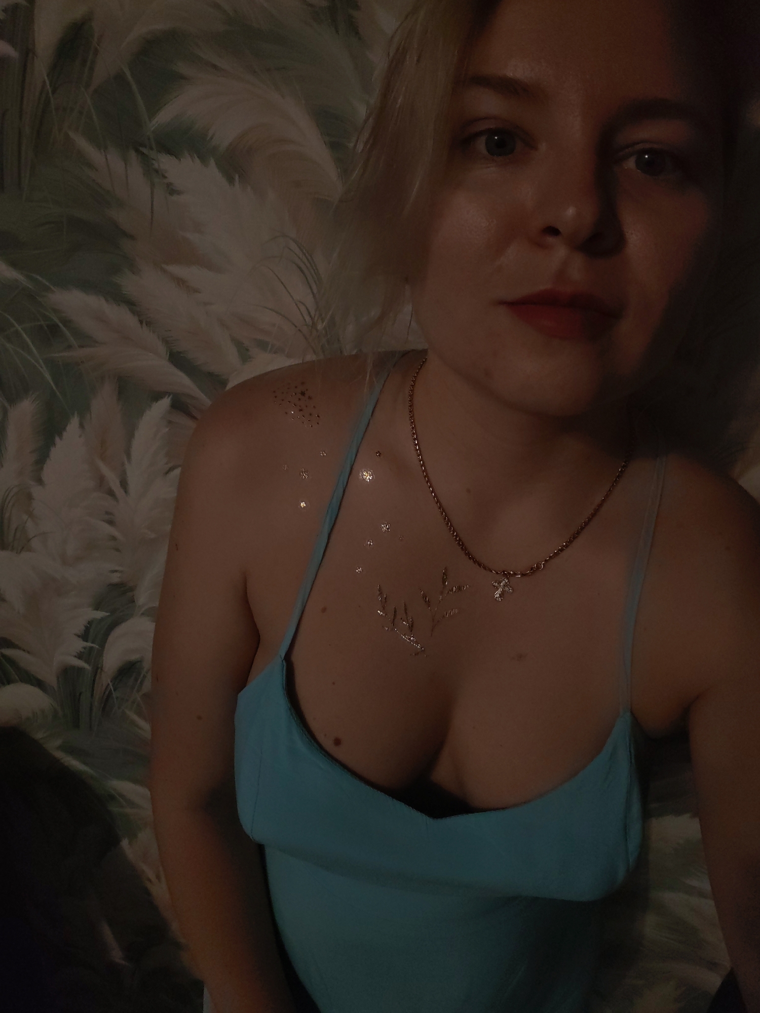 The image shows a medium shot of a woman with blonde hair and light skin, wearing a blue dress and a necklace with a cross pendant. She has a few small metallic star-shaped temporary tattoos on her chest and shoulder. The background appears to be wallpaper with a feathery or grass-like pattern. The lighting is dim, with a spotlight effect on the woman's face and chest. The location is described as Izhevsk, Russia.