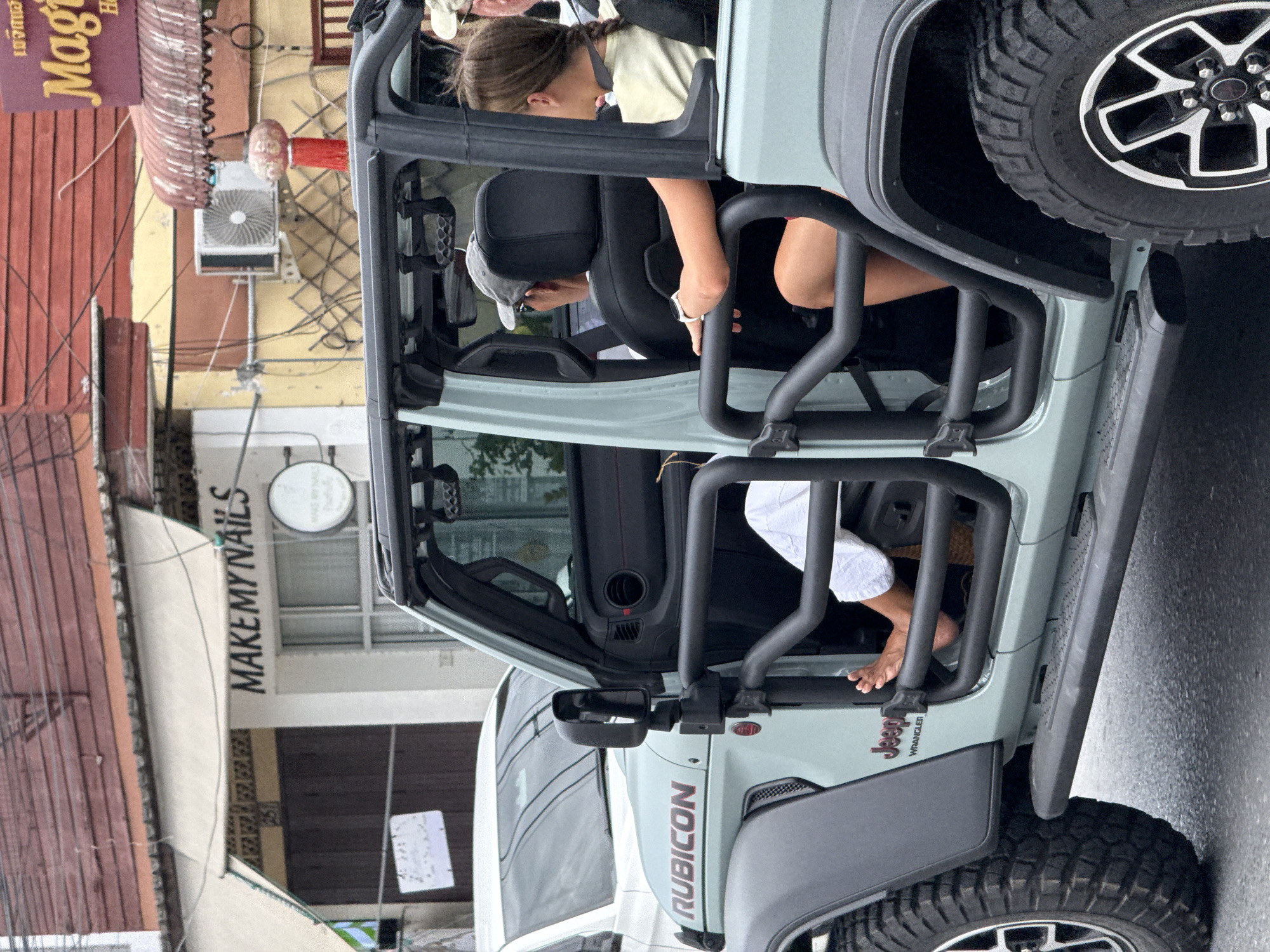 The image captures a street scene in the Ko Samui District of Thailand. In the foreground, a light blue Jeep Wrangler is parked on a wet asphalt road. The Jeep has a roll cage and its doors are open. A person's bare foot is visible extending from the driver's side of the Jeep. Inside the Jeep, two individuals are seated in the back, both facing away from the camera. The person on the left wears a grey baseball cap and a white watch on their left wrist. The person on the right has their hair in a braid.
In the background, buildings line the street. The building to the left has a white facade with a sign that reads "MAKEMYNAILS." A beige awning extends over the storefront. Another building to the right displays a sign with the word "Magic" in large, stylized letters, suggesting a hotel or similar establishment. Various wires and a red lantern are visible on the facade of this building. A white car is partially visible parked behind the Jeep on the left. The overall impression is a typical urban street scene in Thailand.