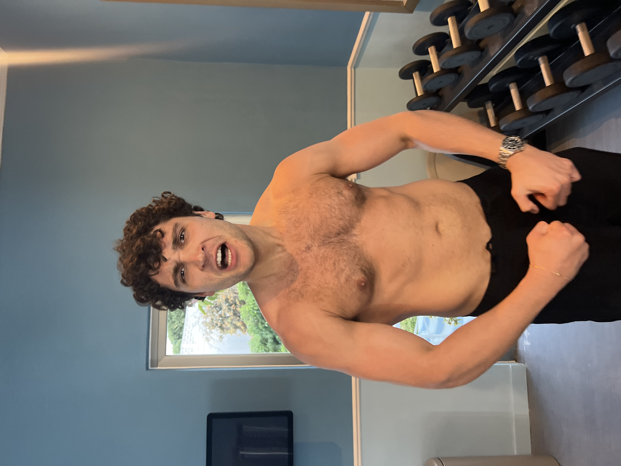 A muscular, shirtless man with curly hair is flexing his biceps in an indoor setting. He is wearing black pants and a watch on his left wrist. In the background, there is a wall painted a light blue color, a window with a view of greenery, and a rack of dumbbells. A black monitor is visible in the lower part of the frame, and there is a cylindrical object near the bottom right corner. The location cannot be determined to be Santa Eulària des Riu, Spain, based on the image provided.