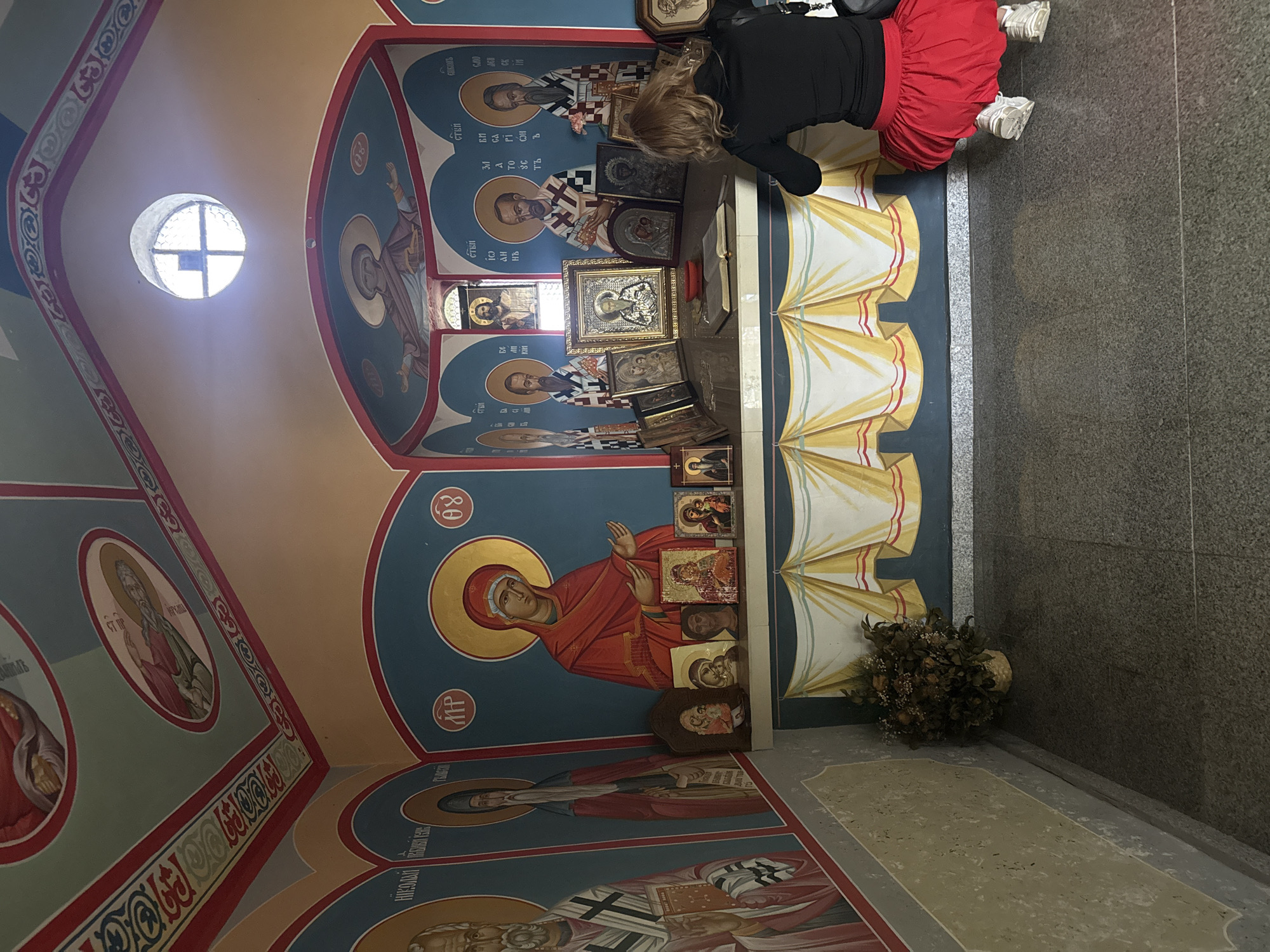 The image shows the interior of a small room, likely a chapel or an icon corner, decorated with religious iconography. The walls and ceiling are painted with religious figures and ornate borders. In the center of the room, a woman with blonde hair, wearing a black top and a red skirt, is crouching down and looking at a display of religious icons on a shelf. The shelf is adorned with a draped fabric that resembles curtains. Various framed icons are arranged on the shelf, some with golden frames. Behind the shelf, a large mural depicts religious figures. To the left of the shelf, there is a basket with dried flowers. The floor is tiled with a speckled granite material. A circular window with a cross-shaped division is visible on the ceiling, letting in light. The overall scene suggests a place of worship, possibly within a church or a private dwelling. Belitsa, Bulgaria is mentioned in the prompt as the location.