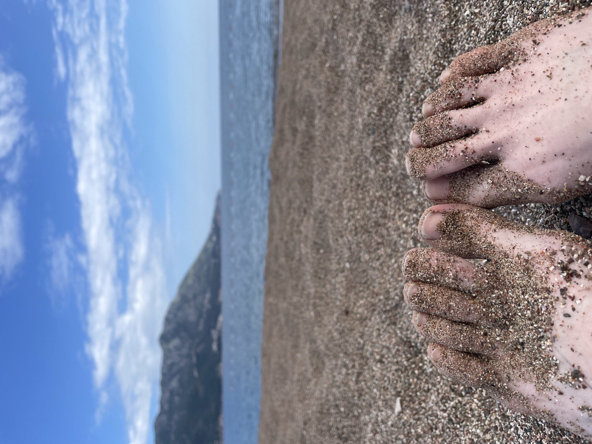The image shows a view of a beach, looking down at two sandy feet. The beach appears to be made of pebbles or coarse sand. The background includes the sea, and a blurred image of a mountain under a partly cloudy sky.
Given the prompt context, the location can be inferred to be Bečići, Montenegro.