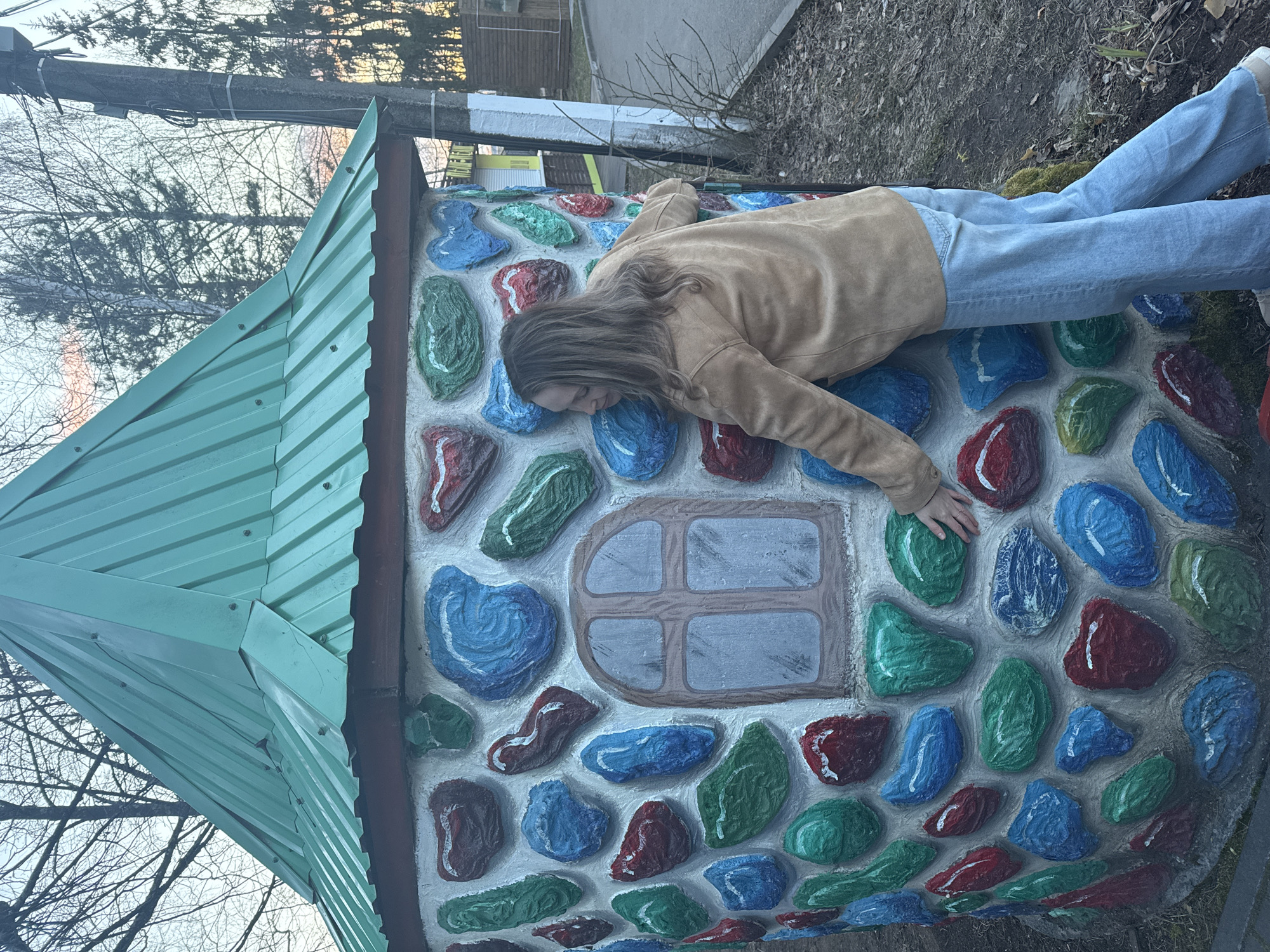 The original image depicts a person lying prone on the side of a whimsical, house-like structure in an outdoor setting. The scene is located in Chaikovsky, Russia.
The primary subject is a person, likely female, with long brown hair, wearing a light brown or tan jacket and light blue jeans. They are positioned horizontally across the decorated wall of the structure, with their head resting against it, turned towards the left. Their arms are outstretched to their sides, hands splayed on the colorful surface. Their posture suggests a relaxed, playful, or contemplative mood, possibly posing for a photograph.
The prominent object is a small, fanciful building, resembling a miniature house. It features a pointed, multi-faceted roof made of light green corrugated metal. Its walls are adorned with large, raised, irregularly shaped elements painted in vibrant blue, green, and red, set against a light gray or white background, creating the appearance of a colorful mosaic or cobblestone pattern. A painted, wooden-framed window with gray panes is visible on the wall, located beneath the person's head.
The setting is outdoors, likely a park or garden. The background reveals bare tree branches against a soft, light sky, suggesting late autumn or early spring. Hints of a utility pole or fence post and dry ground cover are also visible. The lighting appears to be natural daytime, possibly late afternoon or an overcast day, providing soft, diffused light. No visible text is present in the image.