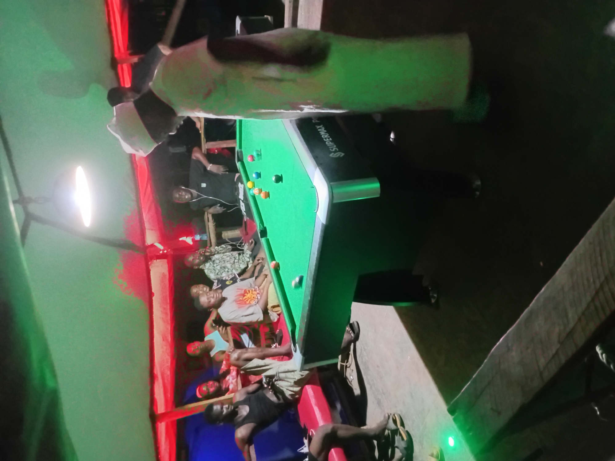 This media file captures a social gathering around a pool table, likely during the evening or night, given the reliance on artificial lighting. The scene is illuminated by a mix of bright white light from a large fixture on the left, and striking red and green colored lights that cast strong hues across the walls and ceiling.
Numerous people, predominantly men, are present. Several individuals are seated on red benches that line the wall, relaxing and observing. Others are standing closer to the pool table, some appearing engaged in the game. One person in a distinctive long white garment and cap stands upright near the top of the frame. Another person in a black tank top and shorts is seated on a red bench in the foreground, with their legs extended.
The central object is a green-felt pool or snooker table with several colored balls scattered across its surface. The brand name "SUPERMAX P" is visible on the side of the table. The floor appears to be made of wood or concrete, with visible wooden beams or structures in the background and foreground, suggesting a somewhat rustic or informal setting. People are dressed casually in t-shirts, tank tops, and sandals. The overall atmosphere appears relaxed and social, indicative of friends or acquaintances enjoying a game and conversation.