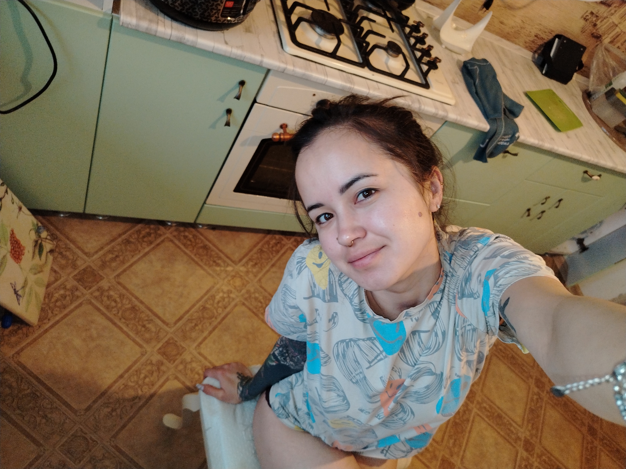 The image shows a person with brown hair in a bun, wearing a patterned t-shirt and visible tattoos on their arm, sitting on a white chair in a kitchen. The kitchen features light green cabinets with bronze-colored handles, a white countertop, and a gas stovetop. A dishtowel and various kitchen objects are on the counter. The floor is covered in patterned brown tiles. The person is taking a selfie. The image does not contain enough information to determine its location within Noviy, Russia.
