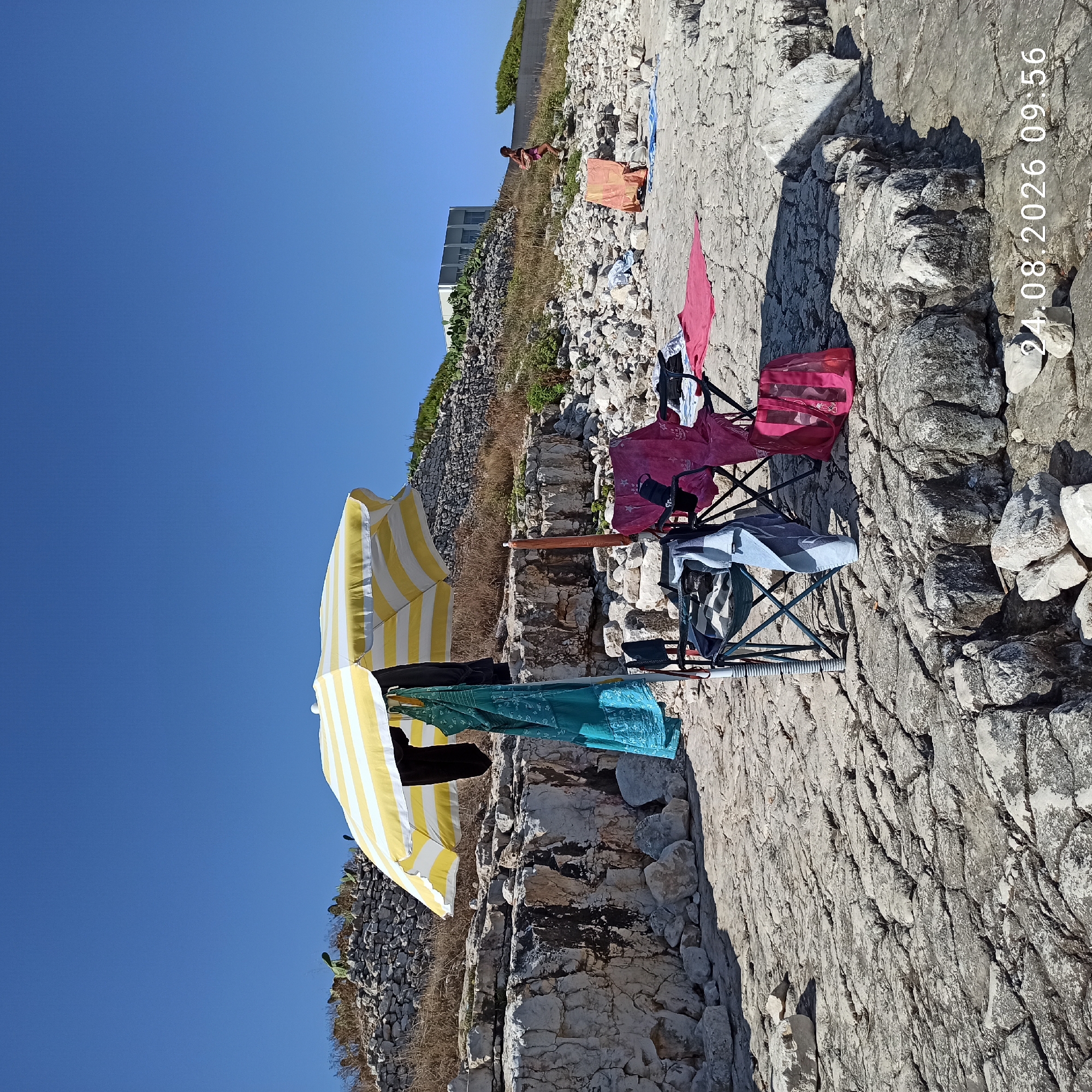 The image depicts a sun-drenched rocky coastline in Molfetta, Italy. A yellow and white striped beach umbrella shades a scene with two folding chairs. Various items of clothing, including a teal dress and a black garment, are draped over the umbrella and a chair. A pink mesh bag rests on the ground near the chairs. A woman in a pink swimsuit is visible walking up a rocky incline in the background. The terrain is characterized by rough, light-colored rocks and sparse vegetation. The sky is clear and blue. A timestamp at the bottom right corner reads "24.08.2026 09:56".