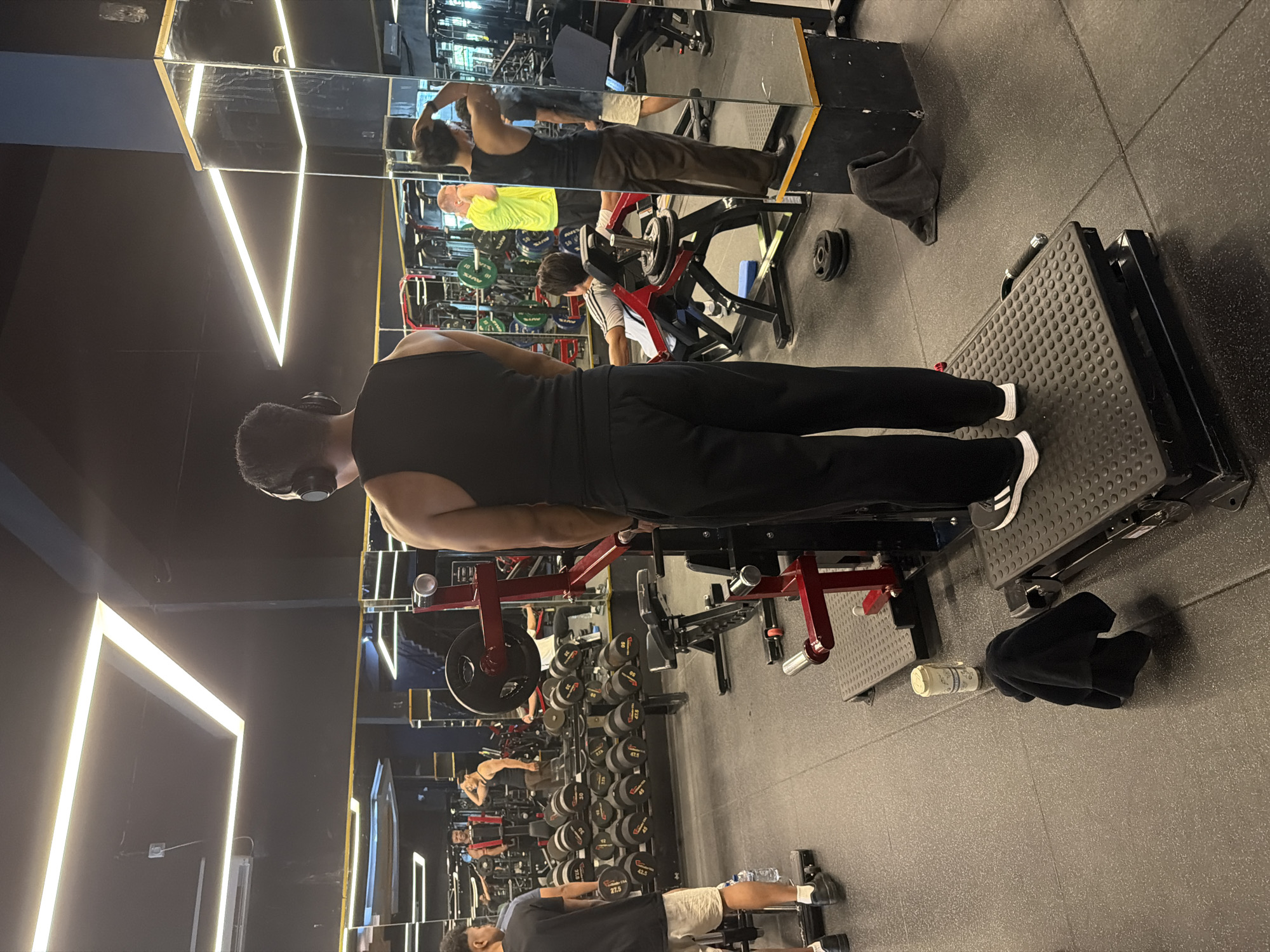 A man wearing a black tank top and black sweatpants, with headphones around his neck and white and black athletic shoes, is seen from behind operating a weight-lifting machine in a gym. The machine is positioned on a black tiled floor with a metallic frame and red supports, featuring a raised, patterned platform for foot placement. To the left, a rack of dumbbells of varying sizes is arranged. A black towel and a clear plastic bottle sit on the floor adjacent to the machine. The background features mirrors reflecting the gym equipment and other individuals exercising. The location is not explicitly known to be Denpasar, Indonesia.