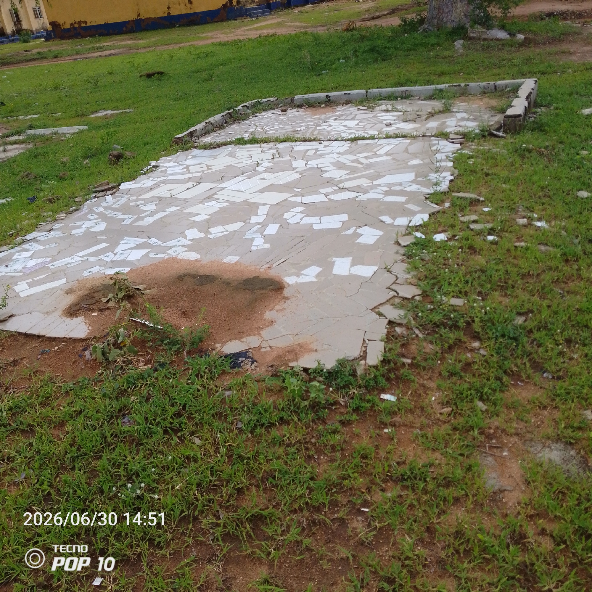 This media file depicts an outdoor scene in Jalingo, Nigeria. The primary subject is a cracked and damaged concrete platform, possibly the foundation of a former structure, tiled with numerous broken white and light gray tiles. The platform is surrounded by overgrown grass and dirt. In the background, a yellow building with a blue trim is visible. The scene appears to be a neglected outdoor area.
There is no discernible activity or event taking place; the scene is static and appears to be a view of abandoned or rundown infrastructure. Notable details include the cracked tiles and the patchy, overgrown vegetation encroaching on the concrete. The lighting suggests it is daytime, but the weather is not clearly indicated.
Visible text in the image includes a timestamp "2026/06/30 14:51" and "TECNO POP 10" indicating the device used to capture the image. The overall impression is one of disrepair and neglect.