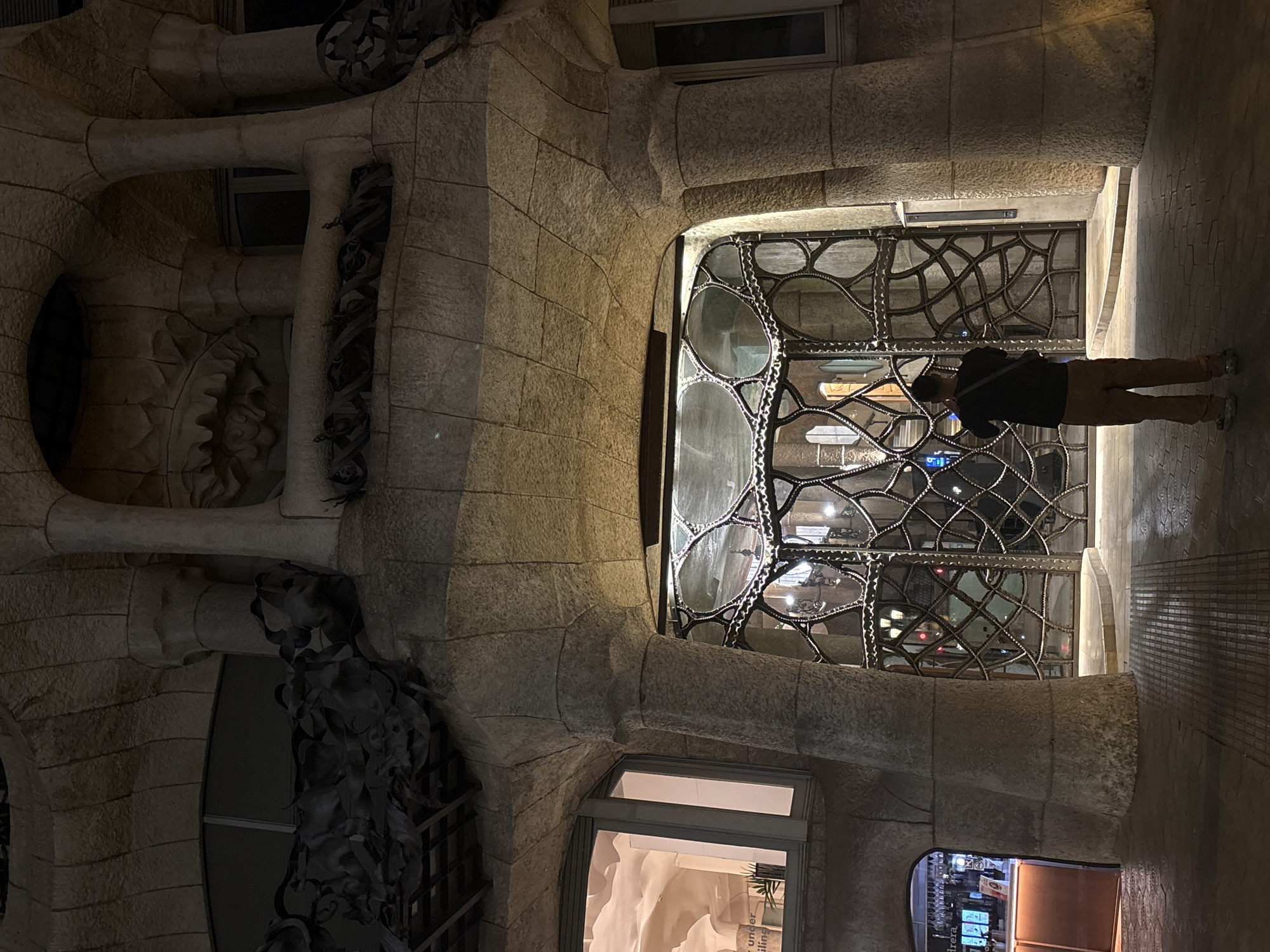 The image shows an exterior view of a building in Barcelona, Spain. The architectural style is characteristic of Antoni Gaudí, possibly Casa Milà (La Pedrera). The building is constructed with light-colored stone and features organic, undulating shapes. A window with an intricate, wrought-iron design is visible. A person, wearing a dark jacket and light-colored pants, is standing in front of the window, possibly taking a photograph. The time of day appears to be evening or night, judging by the artificial lighting emanating from within the building and the darkness of the sky.