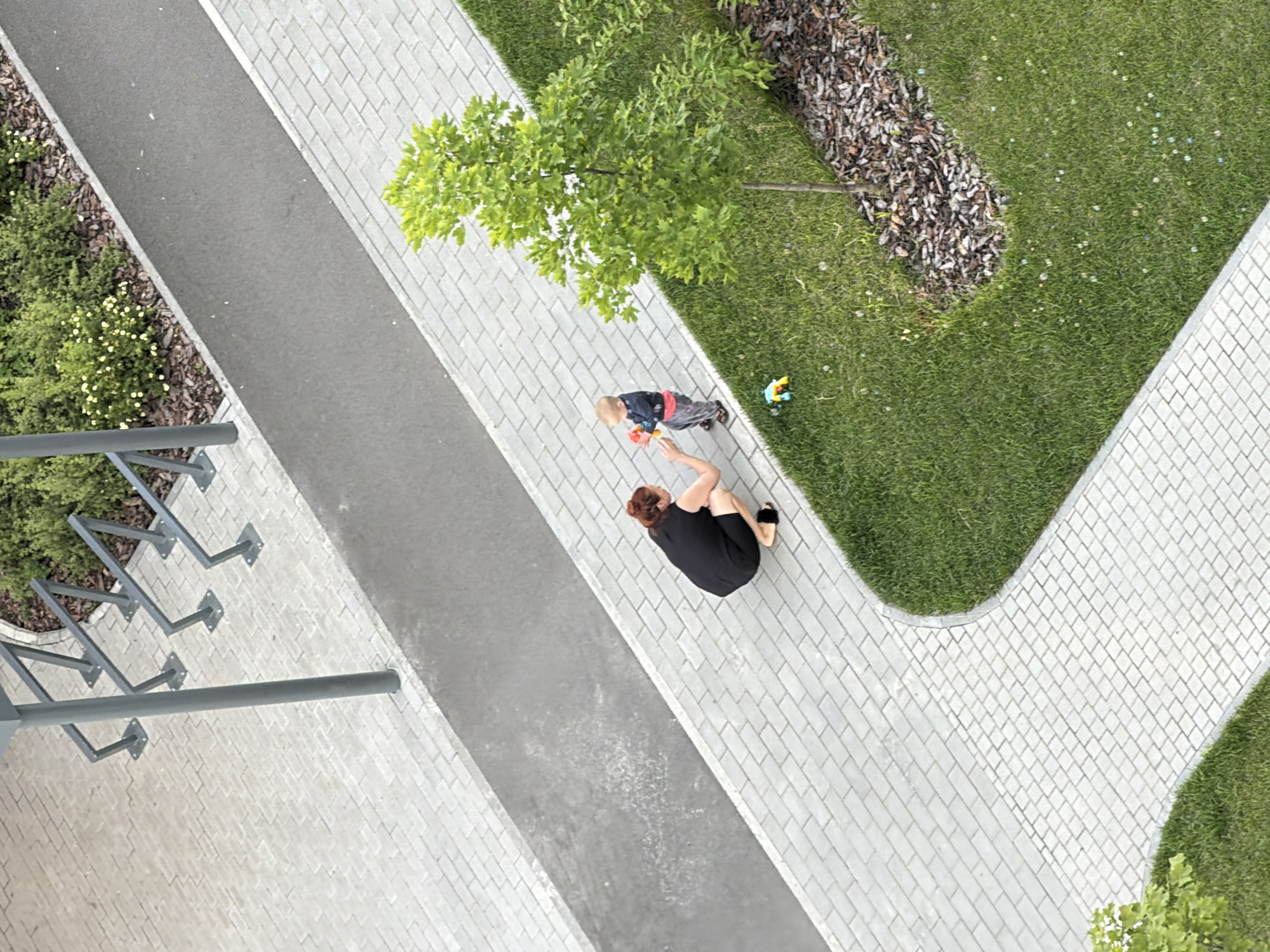 The image depicts a top-down view of a paved walkway in a city environment. A woman, dressed in a black dress and sandals, is crouched down, facing a young child. The child, wearing a dark long-sleeved shirt and grey pants, is standing and appears to be holding a red and yellow toy. The walkway is made of light-colored rectangular pavers. Adjacent to the walkway is a strip of asphalt, and beyond that, a grassy area with a young tree and some bushes. In the background, there is a structure with metal racks, possibly a bike rack. The scene suggests a public outdoor space, like a park or a residential area. The image is sharp and clear, providing a good level of detail of the subjects and their surroundings. There is no information to specify the location within Kazan, Russia.