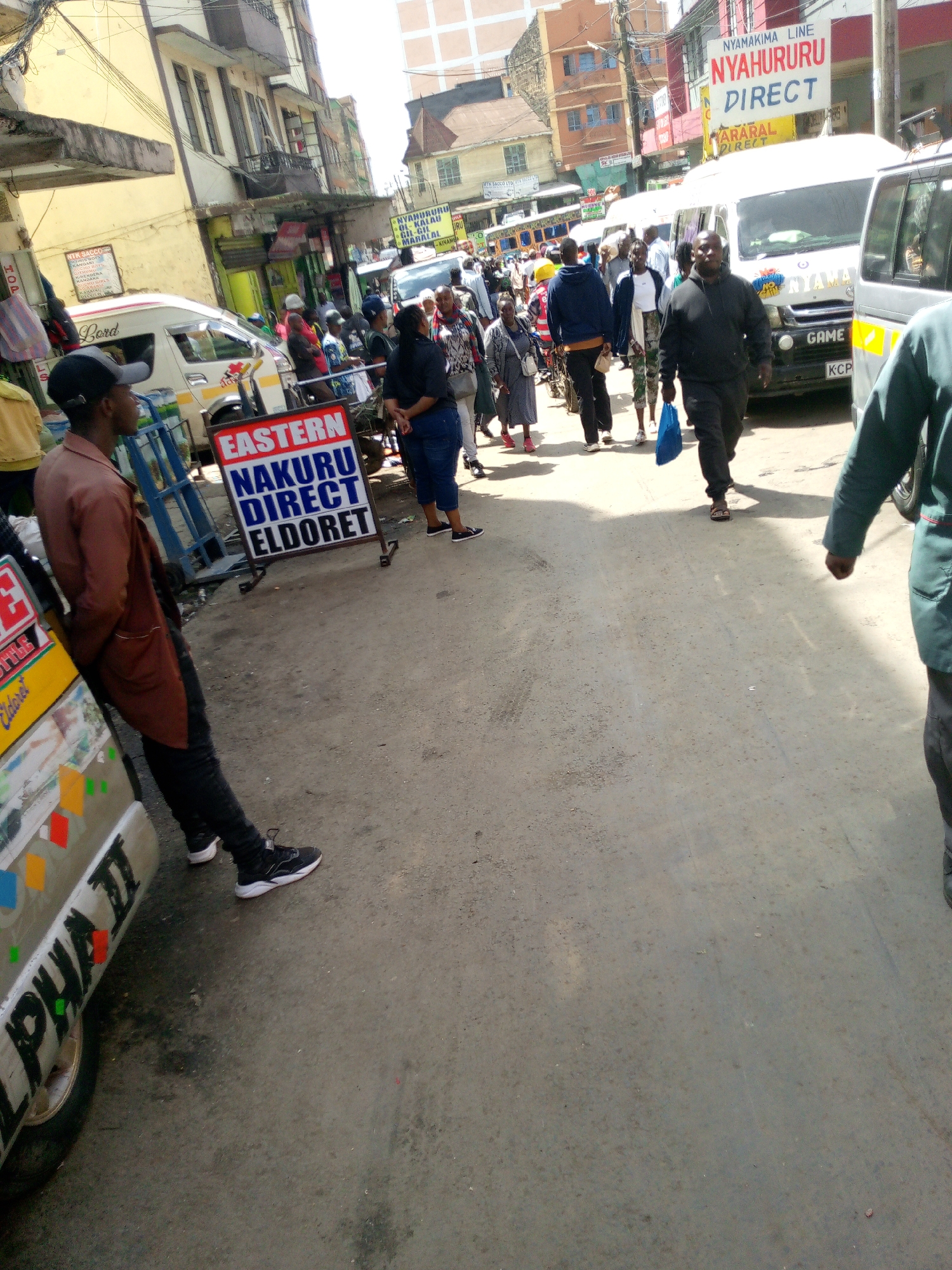 This image captures a bustling street scene in Nairobi, Kenya, specifically within the Nyamakima/River Road commercial area, identifiable by the presence of "NYAMAKIMA LINE" and "NYAHURURU DIRECT" signage, indicative of matatu (public service vehicle) routes. The paved street, a worn gray, is densely populated with approximately 20-30 individuals, mostly adults, engaged in walking and standing. On the left foreground, a male in a black baseball cap, brown long-sleeved shirt, and black pants leans against the front bumper of a white passenger van adorned with colorful decals and text, including "ALPHA II." Adjacent to him, a red and white A-frame sign prominently displays "EASTERN NAKURU DIRECT ELDORET." In the midground, a man wearing a black hoodie and black pants walks towards the camera, carrying a blue bag in his left hand. Directly behind him, a white Toyota Hiace-type matatu is parked or moving slowly, featuring "NYAMA" branding, a "GAME" sticker, and a Kenyan license plate beginning with "KCP." Various other white vans are visible further down the street amidst the pedestrian traffic. Multi-story buildings, characterized by light yellow and brown facades and multiple windows, line both sides of the street under a bright sky. Numerous overhead wires are visible traversing the street between structures. The overall environment suggests a busy urban transportation and commercial hub during daytime.
