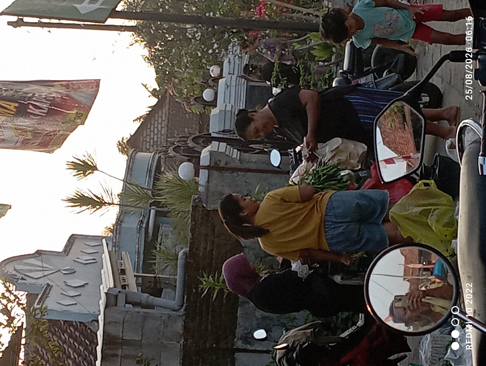 In the city of Dukuhseti, Indonesia, an outdoor scene captures a moment of daily life. Three women and a young girl are present. One woman, wearing a purple hijab and black attire, stands to the left, facing away from the camera and appearing to be engaged in a transaction or conversation with another individual who is not fully visible. To her right, a woman in a yellow t-shirt and denim shorts stands, also looking down, possibly at something in her hands or the ground. A third woman, dressed in a black shirt and striped skirt, is in the center, holding what appears to be cash and interacting with the woman in yellow. A young girl in a teal t-shirt and red shorts stands to the right, looking downwards.
The background features a building with an ornate facade, possibly a house or a shop, with a tiled roof and decorative elements. Vegetation, including palm-like plants and trees, is visible, suggesting a tropical or subtropical environment. A motorcycle is partially visible on the left, and another motorcycle is parked behind the woman in black. A red motorcycle is in the foreground, its side mirror reflecting a person holding a mobile phone. A yellow plastic bag lies on the ground near the motorcycles. The lighting suggests it is either early morning or late afternoon, with the sun casting shadows. The timestamp "25/08/2026 06:16" is visible in the lower right corner, indicating the date and time the photograph was taken.