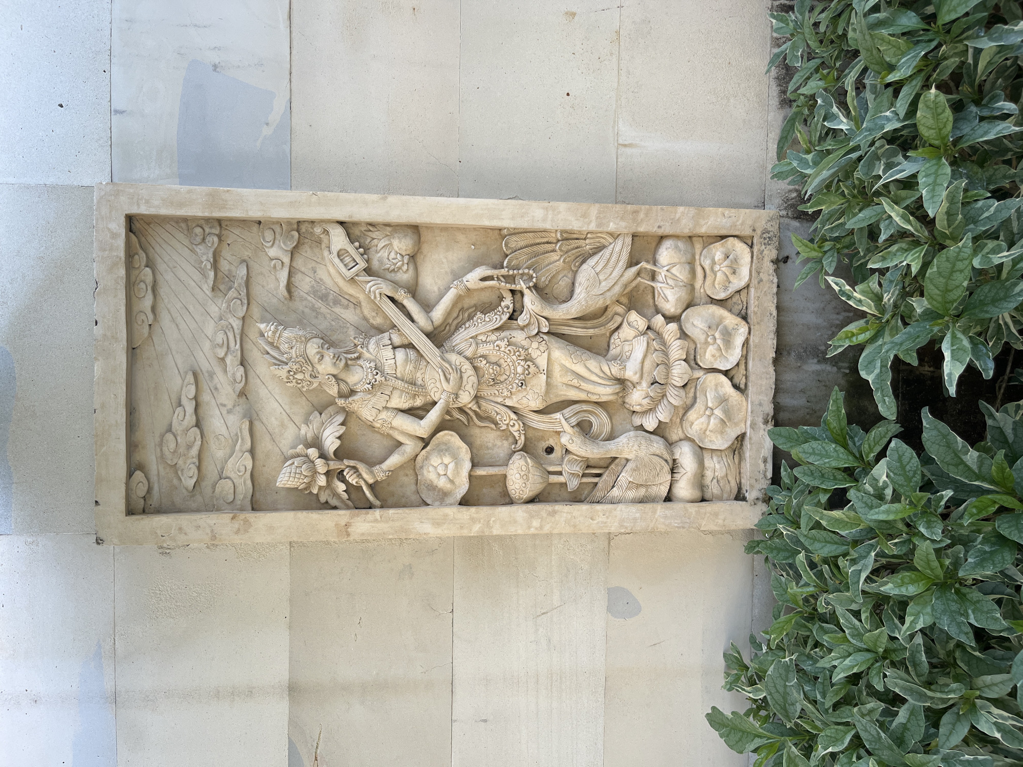 This image features a stone relief sculpture embedded in a light-colored tiled wall. The sculpture depicts a central figure, likely a deity or mythical being, in a reclining or flowing pose. The figure is adorned with intricate jewelry and a headdress. To the left of the figure, a series of stylized clouds and rays suggest a celestial or atmospheric element. To the right, the sculpture incorporates elements of nature, including lotus flowers and a bird with elaborate plumage. The overall style of the carving suggests an Asian, possibly Balinese, influence, consistent with the provided location context of Kuta Selatan, Indonesia. The sculpture is set outdoors, with lush green foliage visible on the right side of the frame, hinting at a garden or courtyard setting. The lighting suggests it is daytime, with soft shadows indicating diffused sunlight. The stone appears aged, with some minor imperfections that add to its character. No visible text is present in the image.