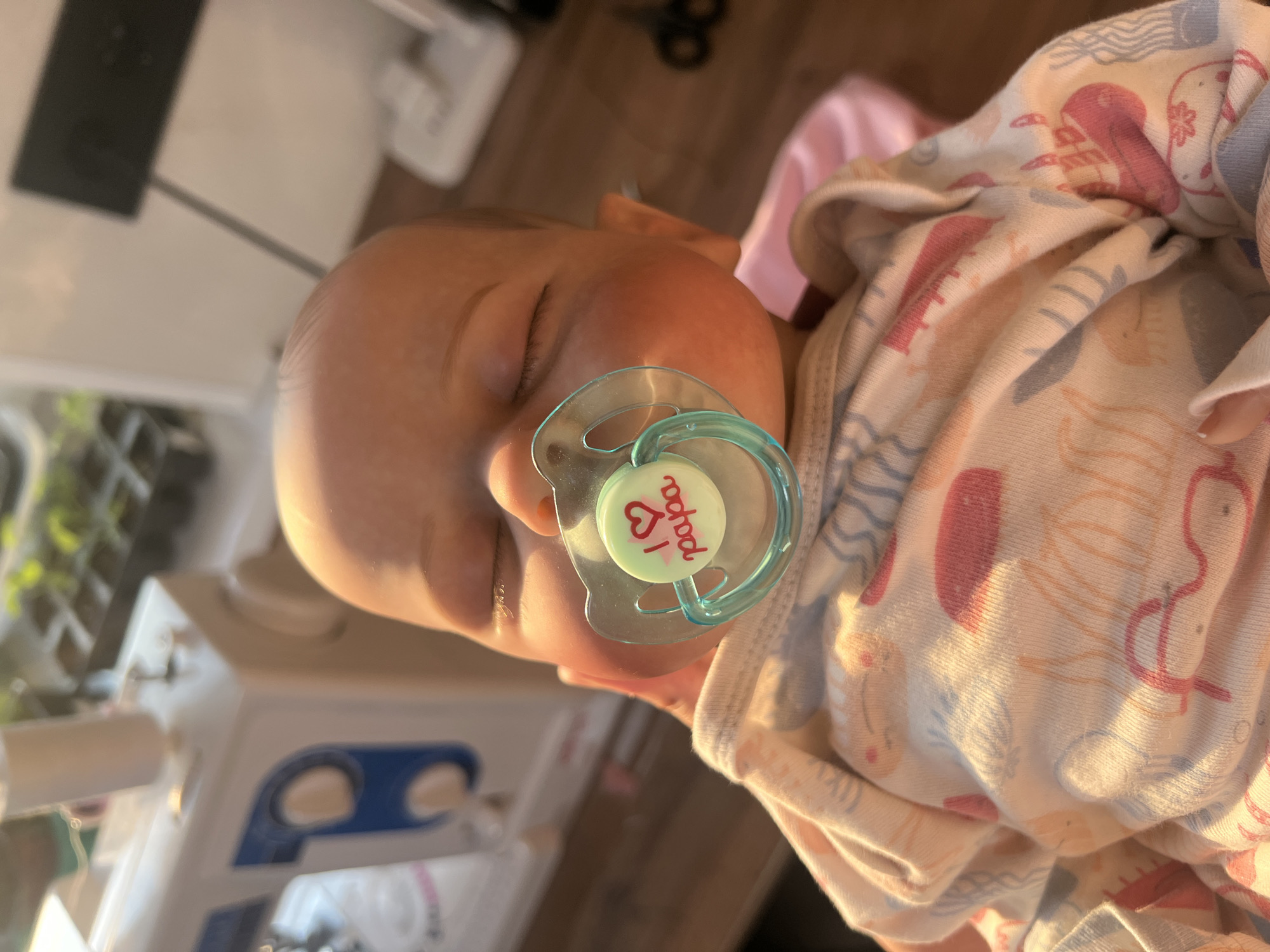 The media file shows a close-up, horizontally oriented image of a sleeping infant. The baby is wearing a light-colored onesie with a pattern of sea creatures like jellyfish and possibly whales in pink and blue. A teal pacifier with the text "I ❤️ papa" is in the baby's mouth. The baby's eyes are closed, suggesting peaceful sleep, and the lighting indicates it might be daytime, possibly late afternoon or early morning due to the warm, soft glow. The background is out of focus but appears to be an indoor setting with a wooden floor and some blurred objects, including what looks like a white electronic device and some greenery. The overall mood is serene and tender. The text "I ❤️ papa" is visible on the pacifier.