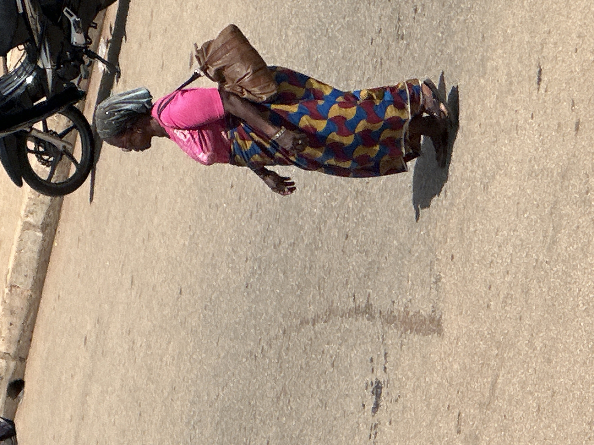 In this full-shot photograph, a person is walking along a paved surface, possibly a road or sidewalk, in Bukuru, Nigeria. The person, a woman, is dressed in a vibrant, colorful patterned skirt and a bright pink top. She is carrying a brown bag over her shoulder. Her head is covered with a dark, possibly fabric, wrap. Her skin tone suggests she is of African descent.
The scene appears to be outdoors during the day, as indicated by the clear shadows cast on the ground. The lighting suggests it's a sunny day, possibly midday. The ground is a textured, light-colored pavement. To the left of the woman, part of a black motorcycle is visible, including its rear wheel and fender.
The woman's posture and the way she is walking suggest she is on a journey or moving from one place to another. Her facial expression is not clearly discernible. The overall impression is of a daily scene in an urban or semi-urban environment in Nigeria. There is no visible text in the image.