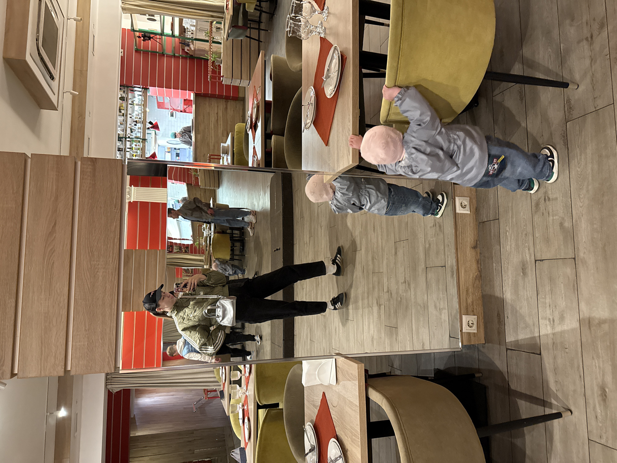 A vertically oriented photograph shows the interior of a restaurant in Moscow, Russia, with a large mirror reflecting the scene and people in it. In the foreground, there is a table set for dining with plates, cutlery, and glasses. Adjacent to the table, a young child is playing with a chair, wearing a light blue jacket, jeans, and sneakers, with a pink hat covering the head. The mirror reflects the child's image.
In the mirror's reflection, a person wearing a black cap, an olive green jacket, black pants, and black sneakers with white socks is visible taking a picture with a mobile phone. A silver purse hangs from the person's shoulder. The reflection shows the person standing in the restaurant. The mirror also reflects other patrons in the restaurant, with red-striped decor visible in the background, and glimpses of a bar with bottles.
The restaurant interior has wooden flooring and the furniture consists of wooden tables and cushioned chairs. The lighting appears to be ambient.