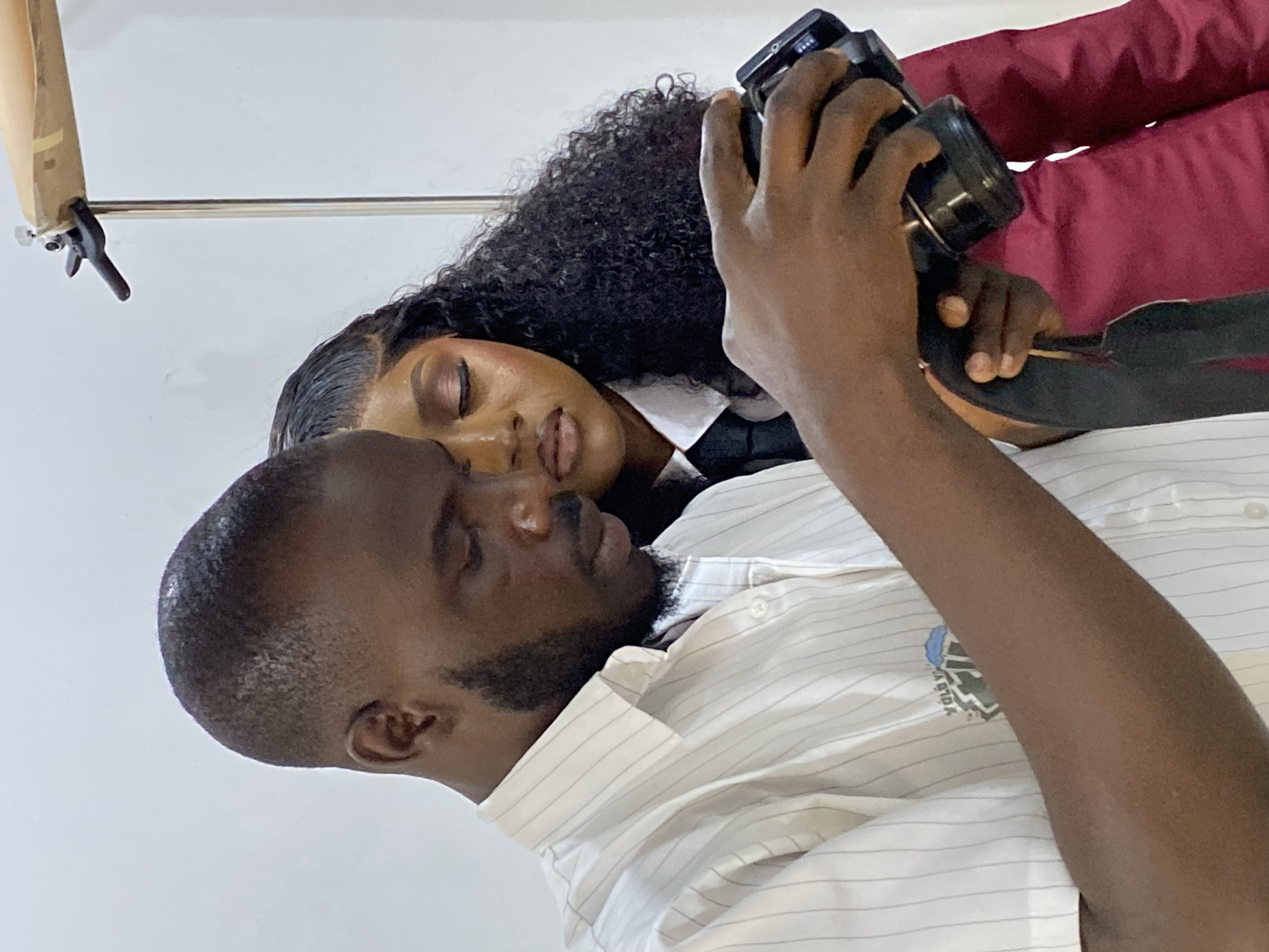 This image captures two individuals, a man and a woman, in an indoor photography setting in Gura Top, Nigeria. The background consists of a plain white wall. In the upper right corner, a metal stand with a black clamp and a beige, fabric-wrapped object is visible, indicating studio equipment.
The man, positioned in the foreground and to the left, is dark-skinned with short dark hair and a trimmed beard. He is wearing a white collared shirt with thin grey vertical stripes; a faint blue and green emblem is visible on his right bicep area. He holds a black digital camera with both hands, looking downwards at its screen or controls, with a black strap around his neck.
Behind and to the right of the man stands a dark-skinned woman with long, dark, curly hair. She is dressed in a white collared shirt, a black tie, and a maroon/burgundy jacket. Her eyes are directed downwards, appearing to observe the camera or the man's actions. Both individuals are focused on the camera, suggesting a shared activity involving photographic review.