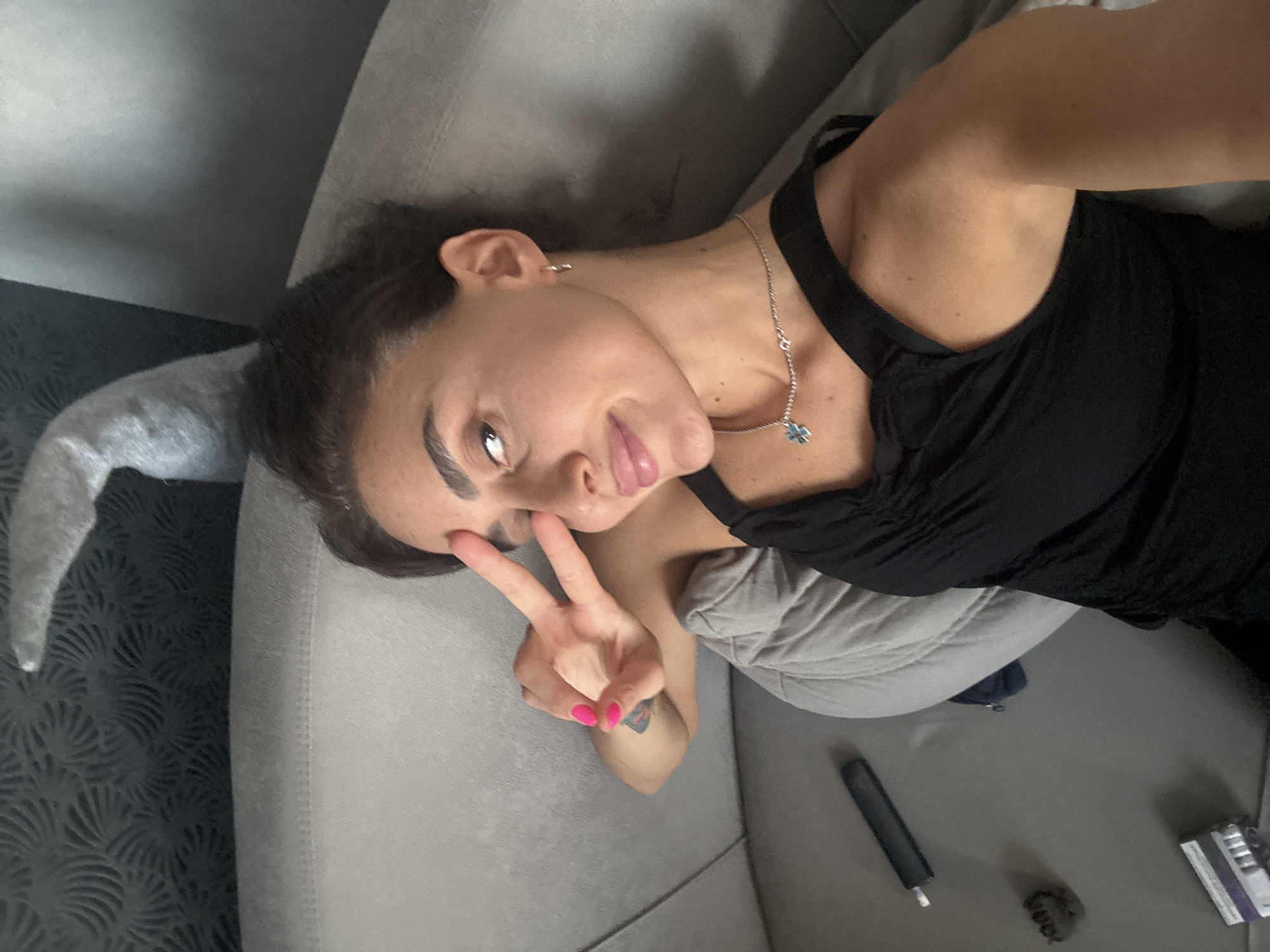 A young woman with dark hair and dark eyebrows, wearing a black tank top and a silver chain with a blue pendant, is lying on a sofa and holding up two fingers in a peace sign. Her right arm is bent and positioned in front of her face, obscuring part of it. She is looking at the camera with her eyes slightly narrowed and her mouth slightly open, with full lips. Her nails are painted bright pink. A pillow is visible behind her head, and a dark patterned wallpaper is in the background. To the left of the sofa, a black electronic device and what appears to be a pack of cigarettes are visible on a flat surface. The scene appears to be indoors, likely in a residential setting.