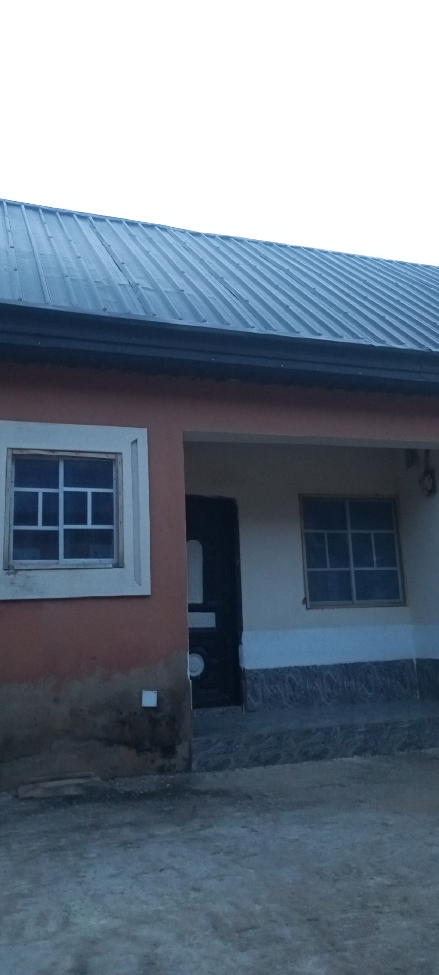 The image shows the exterior of a single-story building with a dark gray corrugated metal roof. The building has a light peach-colored stucco exterior. On the left side of the building, there is a square window with a white frame, divided into smaller panes by a grid pattern. Below the window, the stucco is stained and appears weathered.
To the right of the window is an entrance with a dark door that has a decorative white panel. The entrance is recessed and has a lighter-colored ceiling. To the right of the entrance, there is another square window with a similar grid pattern. The lower portion of the building, below the windows and entrance, is decorated with a patterned tile that appears to be black and gray.
The ground in front of the building is a rough, textured surface, possibly concrete or gravel, with some debris. The sky above is overcast and appears bright white. The overall impression is that of a simple, possibly residential or commercial building in an urban or semi-urban environment.