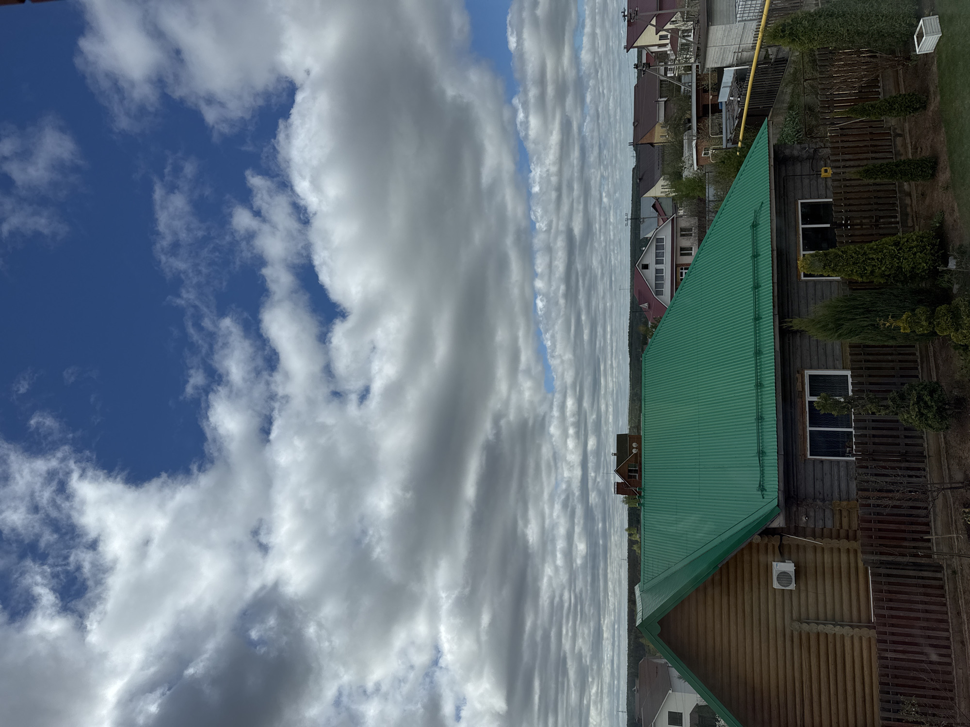 The image depicts a view in Прикамский, Russia. The foreground features a wooden house with a green corrugated metal roof. Adjacent to the house is a wooden fence and some shrubbery. Beyond the house and fence are other houses in the neighborhood. The sky occupies a significant portion of the image, exhibiting a combination of blue sky and layered, striated clouds. The photograph is taken from a slightly elevated position, providing a broad view of the residential area and the sky above.