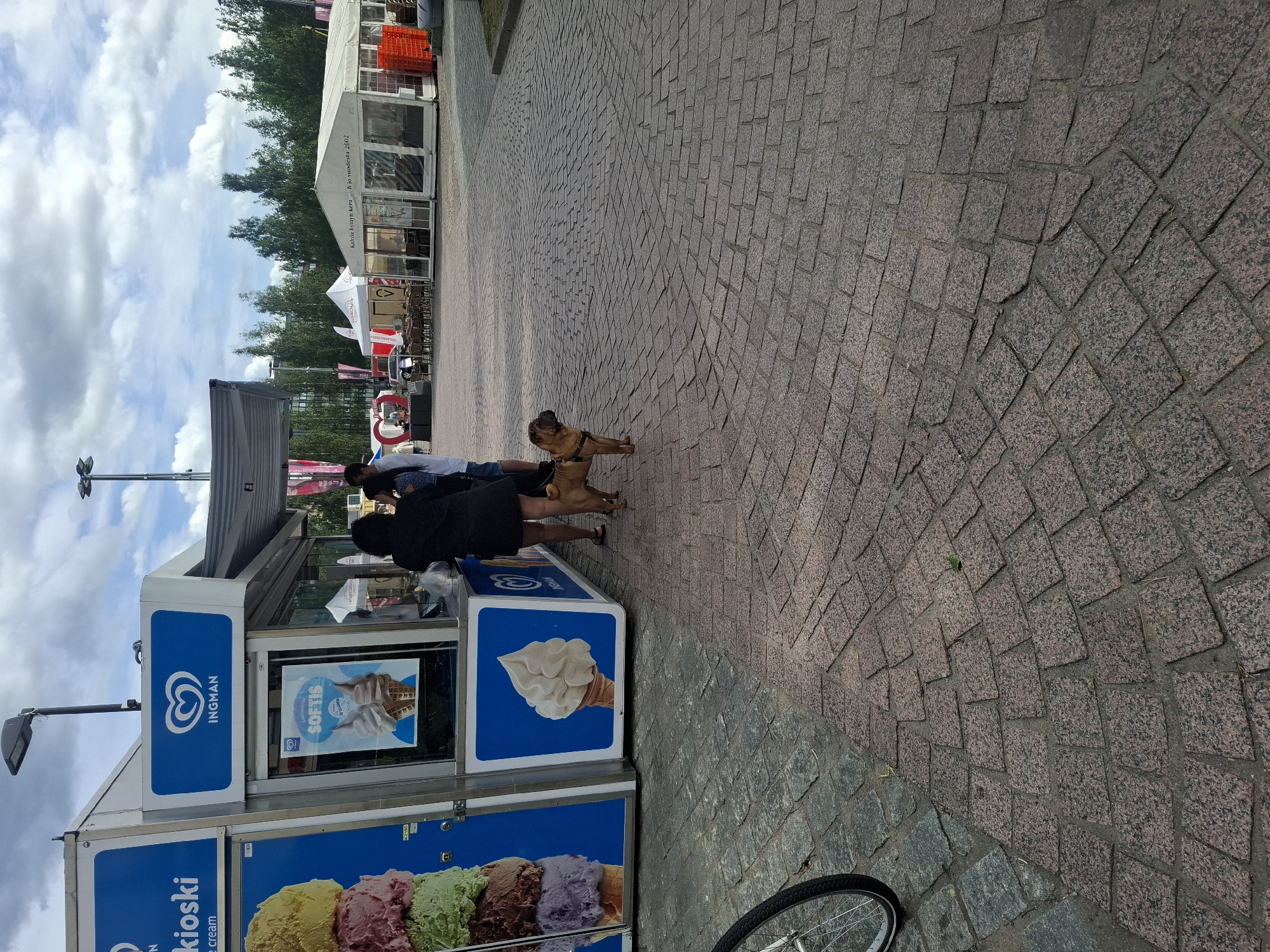 The image depicts an outdoor scene at an event or market in Mikkeli, Finland, with an Ingman ice cream kiosk in the foreground. Two adults, one female wearing a black dress and another partially visible, are standing near the kiosk, with the female in the black dress interacting with a small, tan-colored dog on a leash. The dog is facing away from the camera. The kiosk has a prominent Ingman logo and displays pictures of ice cream. The ground is paved with cobblestones. In the background, several white tents are set up, suggesting a festival or fair. Trees line the horizon under a cloudy sky. A bicycle wheel is visible in the lower left corner of the image.