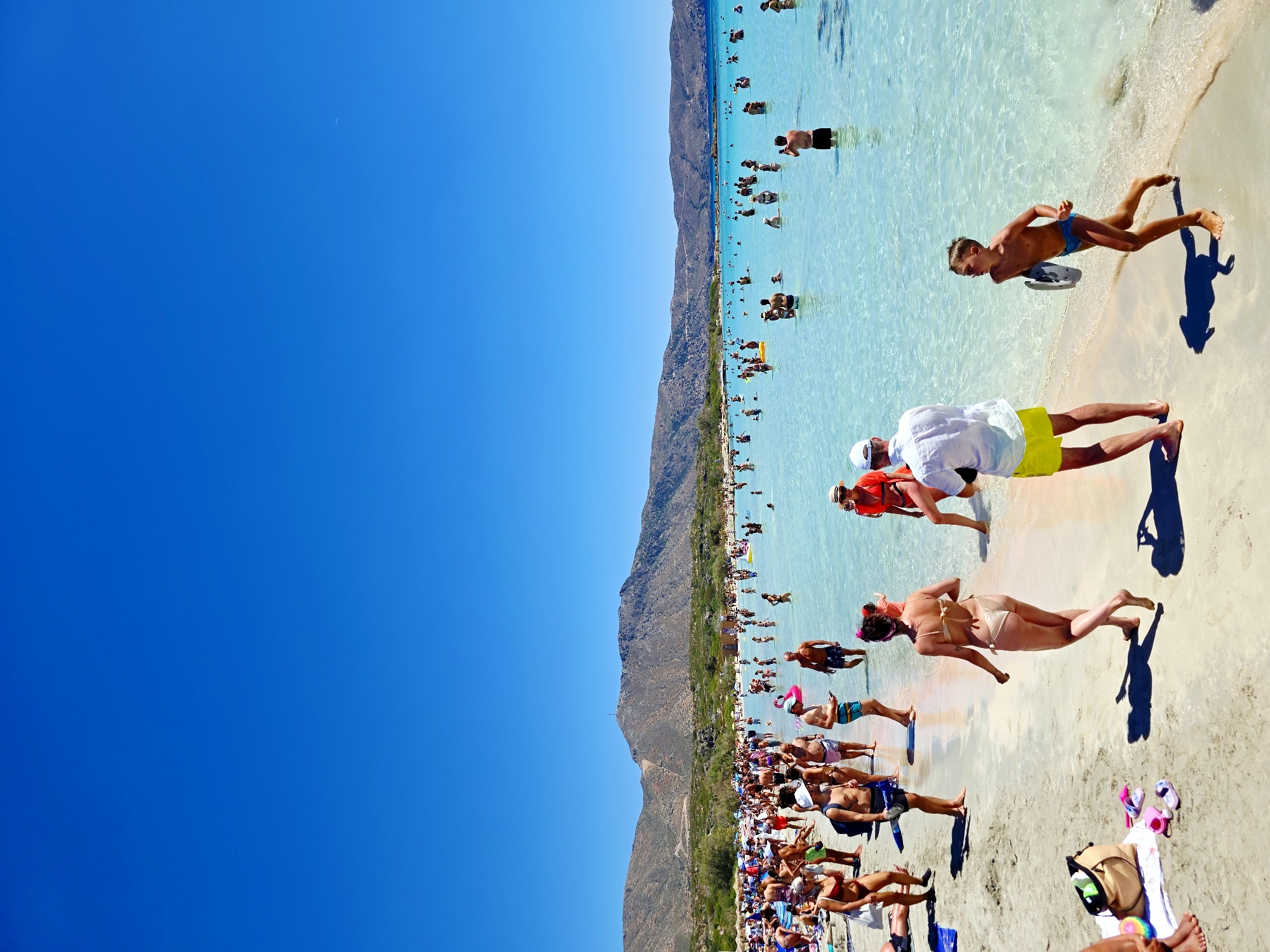 This image depicts a vibrant beach scene on a sunny day, likely in Greece, given the location context. The turquoise water is shallow and clear, with many people enjoying the ocean. The beach is crowded with sunbathers lounging on the sand and others wading in the water. A backdrop of rugged, sun-drenched mountains rises behind the beach, adding to the scenic beauty of the location.
The activity is primarily leisure and recreation. People are swimming, walking along the shore, and relaxing under the bright sun. The mood appears to be cheerful and relaxed, with individuals and groups enjoying a day at the beach. The time of day is clearly daytime, with strong sunlight casting distinct shadows, suggesting it's likely midday or early afternoon. The weather is warm and clear, ideal for a beach outing.
Notable details include a variety of beachwear worn by the people, ranging from bikinis and swim trunks to casual attire. Some individuals are carrying beach bags or have towels spread out on the sand. The water's clarity allows for a view of the sandy bottom in many areas. There is no visible text in the image. The overall impression is one of a lively and popular tourist destination on a beautiful summer day.