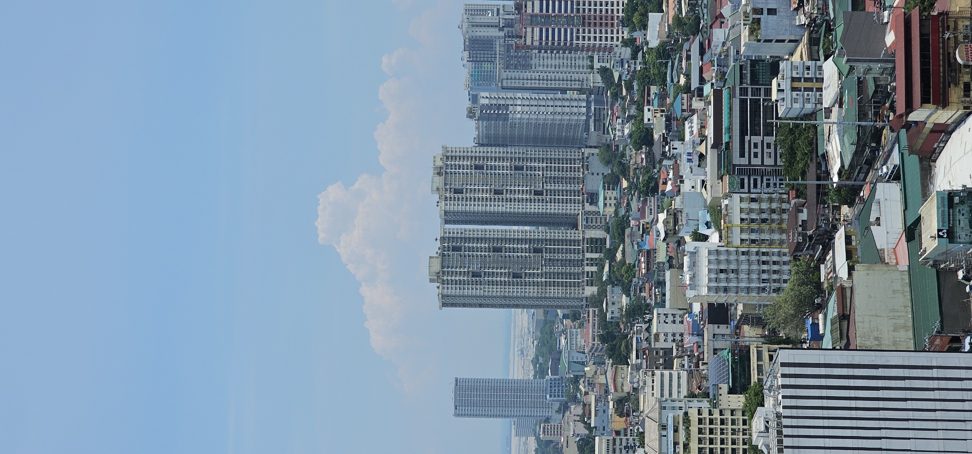 This is a vertical, wide-angle, aerial shot of the city of Makati, Philippines, on a partly cloudy day. The scene is dominated by a dense urban landscape with numerous buildings of varying heights, from mid-rise to skyscrapers. In the background, a tall, slender, white skyscraper stands apart, with two massive, gray residential towers situated to its right. These towers appear to be under construction or recently completed, as some units appear unfinished. Scattered amongst the larger buildings are many smaller, older residential and commercial structures, characterized by diverse architectural styles, colors, and roof types, many with visible signs of wear. The sky is a pale blue with a few large, fluffy white clouds. The overall impression is one of a bustling, densely populated metropolitan area.