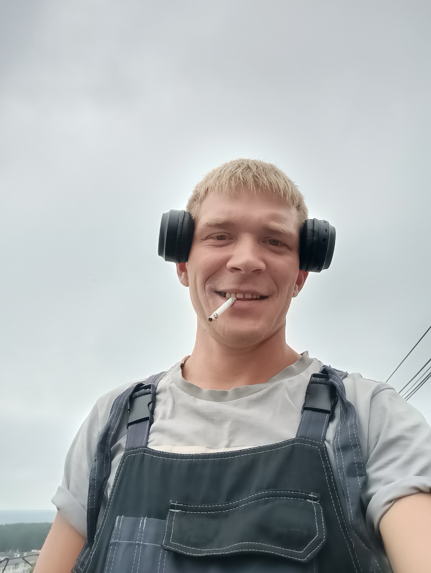 A young man with blond hair and a fair complexion is photographed from the chest up. He is wearing black headphones, and a lit cigarette is in his mouth. He has a gray t-shirt on and dark blue overalls. The man is smiling at the camera. The background is a hazy, overcast sky. The man is outdoors, with what appears to be distant trees and buildings visible at the bottom left of the frame, suggesting a non-urban environment.