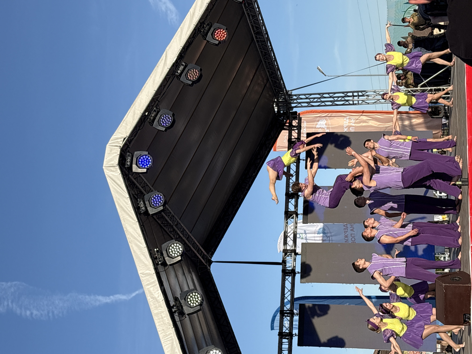 The image shows a performance on an outdoor stage in Khvalynsk, Russia. The stage has a black angled roof with lighting fixtures attached. Below the roof, there is a metal truss structure. A group of performers in purple and yellow costumes are engaged in acrobatic and dance movements on the stage. Some performers are lifting others, creating elevated poses. Behind the performers are flags and screens with text in Russian. The sky is blue and clear in the background.