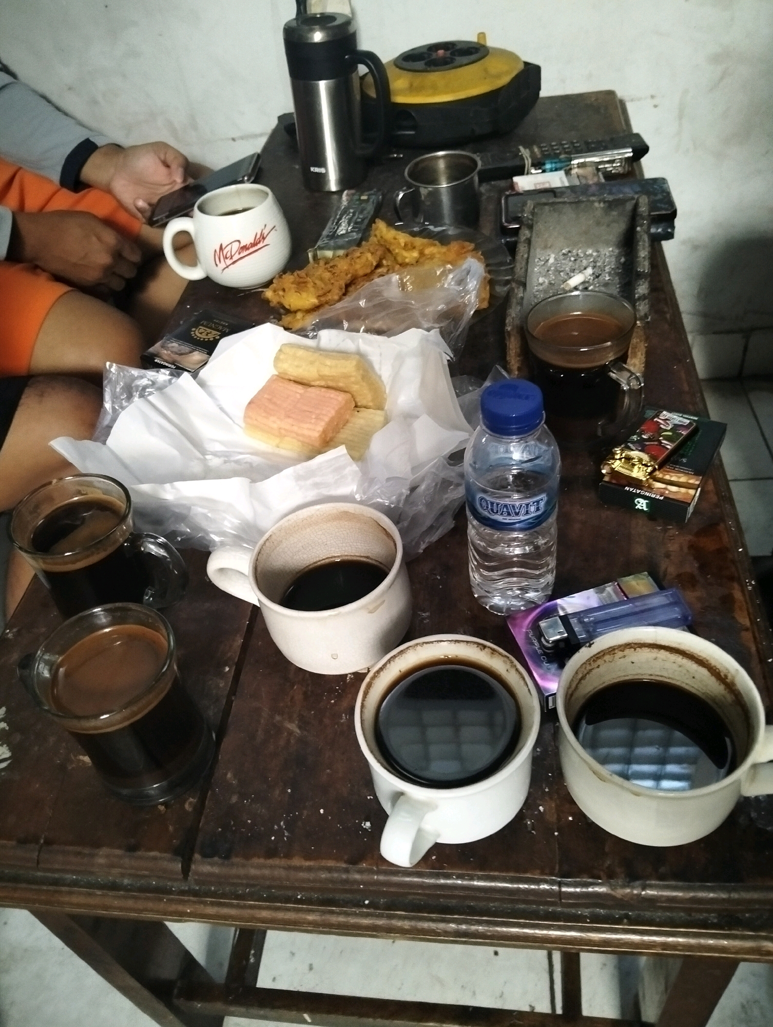 The image depicts a close-up, high-angle shot of a wooden table with various items arranged on it, suggesting a casual gathering or break. The scene appears to be indoors, possibly in a residential or informal commercial setting, in South Jakarta, Indonesia, as indicated by the presence of local cigarette brands and general atmosphere.
In the foreground, four cups of coffee are prominently displayed. Three are white ceramic mugs, one of which is mostly empty with residue on the sides, while the other two are full of dark coffee. A fourth cup, made of clear glass, also contains dark coffee. To the left, a person's arm and hand are visible, holding a smartphone and interacting with it. They are wearing an orange shirt and a grey long-sleeved shirt.
Further back on the table, a white mug with the "McDonald's" logo is filled with coffee. Next to it, a plastic bag contains fried food items, possibly snacks. A metallic thermal flask and a silver metal mug are also present. In the middle of the table, a traditional Indonesian cigarette-rolling tray with used cigarette butts and ash is visible, along with a pack of "Magnum" cigarettes and a lighter. A plastic bottle of "Quavit" mineral water is also on the table. In the background, a yellow and black electronic device, possibly a small appliance or speaker, is partially visible. The overall lighting is dim, creating a somewhat shadowy ambiance. The table surface is worn and shows signs of use, with some debris scattered on it.