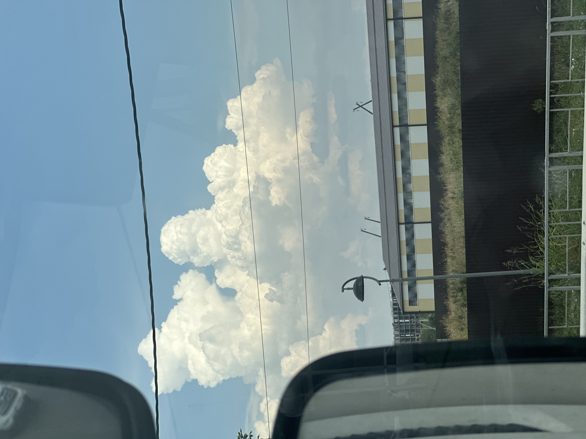 This image captures a view through a vehicle's side mirror and windshield. In the foreground, the dark, curved frame of the side mirror is visible on the left. The scene beyond the mirror is dominated by a vast blue sky with dramatic, billowing cumulus clouds. A dark power line stretches horizontally across the upper portion of the frame. In the middle ground, a long, low building with a grey roof and a facade of alternating white and yellow panels is visible. A dark fence runs in front of this building, separating it from a grassy area. A tall, thin lamppost with a curved arm and a dark lamp is positioned in front of the building and fence. The sky and clouds suggest an outdoor environment, possibly on the outskirts of a city given the presence of the building and power lines. There are no people or discernible actions taking place. The lighting suggests daylight. The image is a static, observational perspective. This scene is likely located in the city of Krasnodar, Russia.