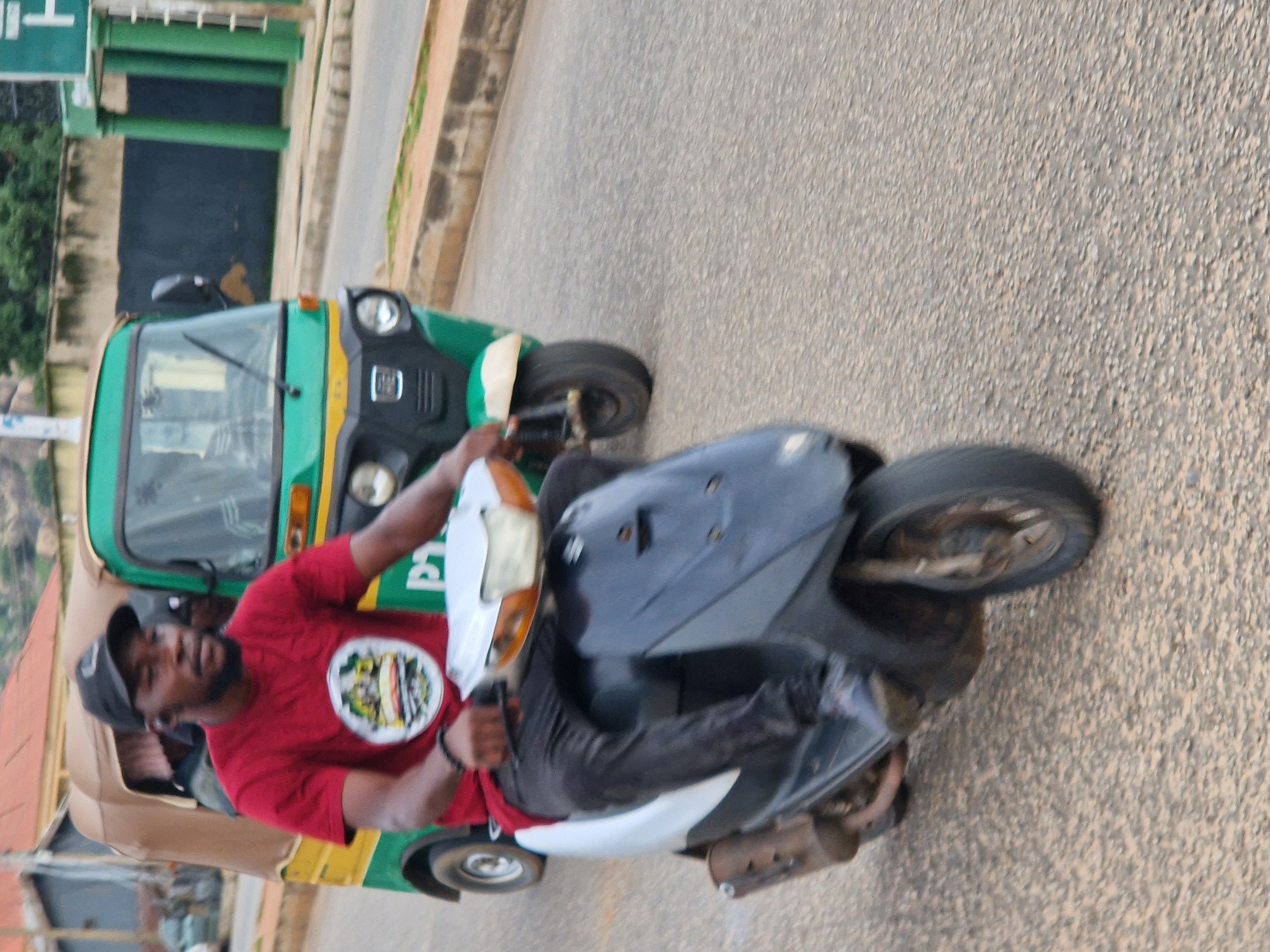 A man wearing a red t-shirt and a black baseball cap rides a scooter on a paved road in Kabong, Nigeria. He has a dark beard and is wearing black pants. He is holding onto the handlebars of the scooter, which is white and black. Behind him, a green and yellow auto-rickshaw is also visible. The auto-rickshaw has a brown canopy. The road is asphalt and appears to be in a residential or commercial area, with buildings and trees in the background. The lighting suggests it is daytime.