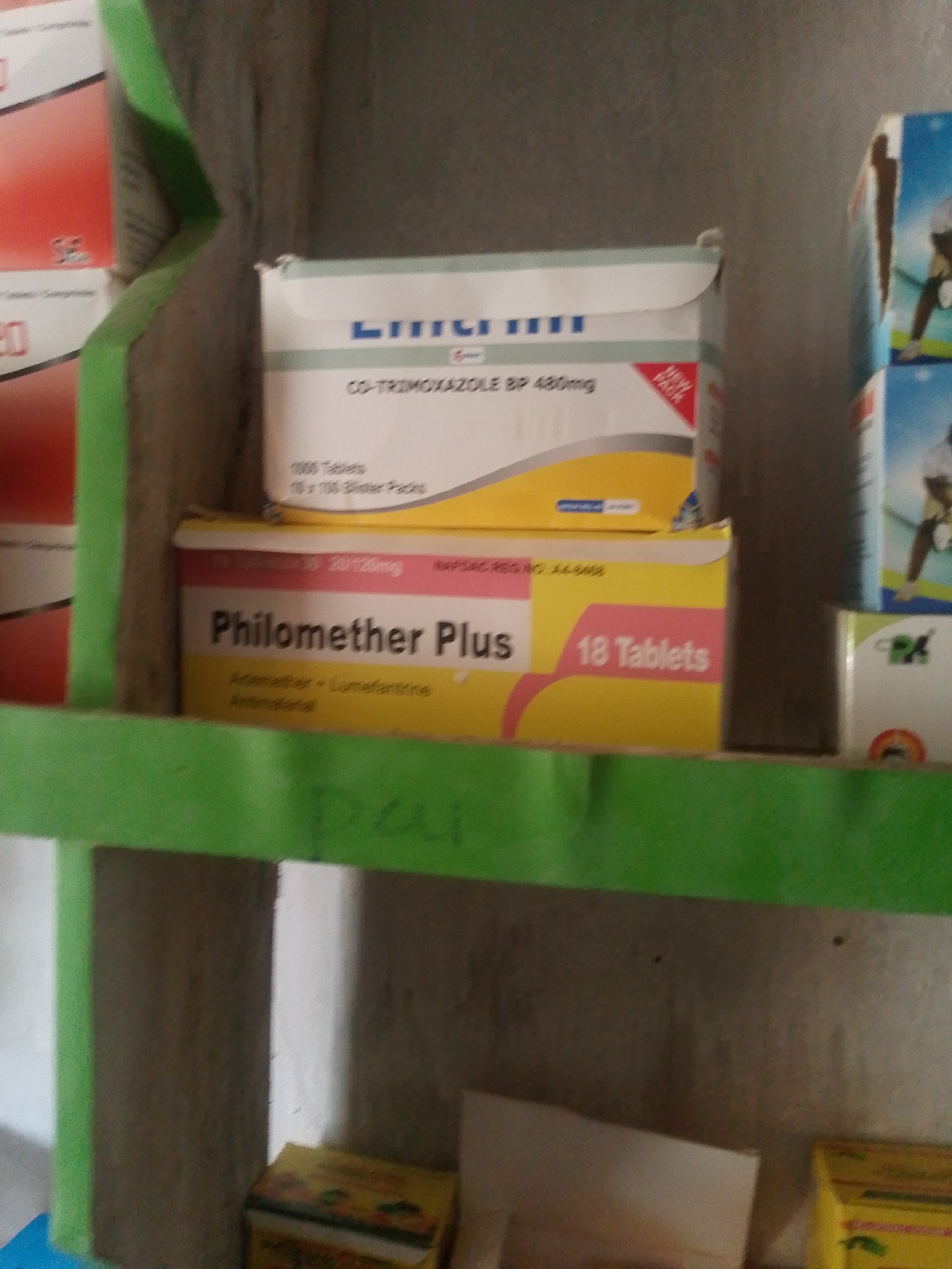 This image depicts a close-up view of shelves in what appears to be a pharmacy or medical supply store. The shelves are stocked with various pharmaceutical products, primarily boxes of medication.
The prominent items on display are two boxes of medicine. The top box, white with yellow and blue accents, is labeled "CO-TRIMOXAZOLE BP 480mg" and contains "1000 Tablets" in "10 x 100 Blister Packs." The text "NEW PACK" is also visible on this box. Below it, a yellow box with pink accents is labeled "Philomether Plus" and indicates "18 Tablets." Beneath this, text reads "Artemether + Lumefantrine Antimalarial."
Other partially visible boxes and items suggest a larger inventory. The background is a textured, neutral-colored wall. The shelves themselves are painted a bright green. The overall scene is one of organized retail display for medical supplies. The lighting appears to be indoor artificial light, with no clear indication of time of day or weather. There are no people visible in the frame. The location context provided is Jalingo, Nigeria, which suggests this is likely a local pharmacy. The visible text is primarily product information and branding.