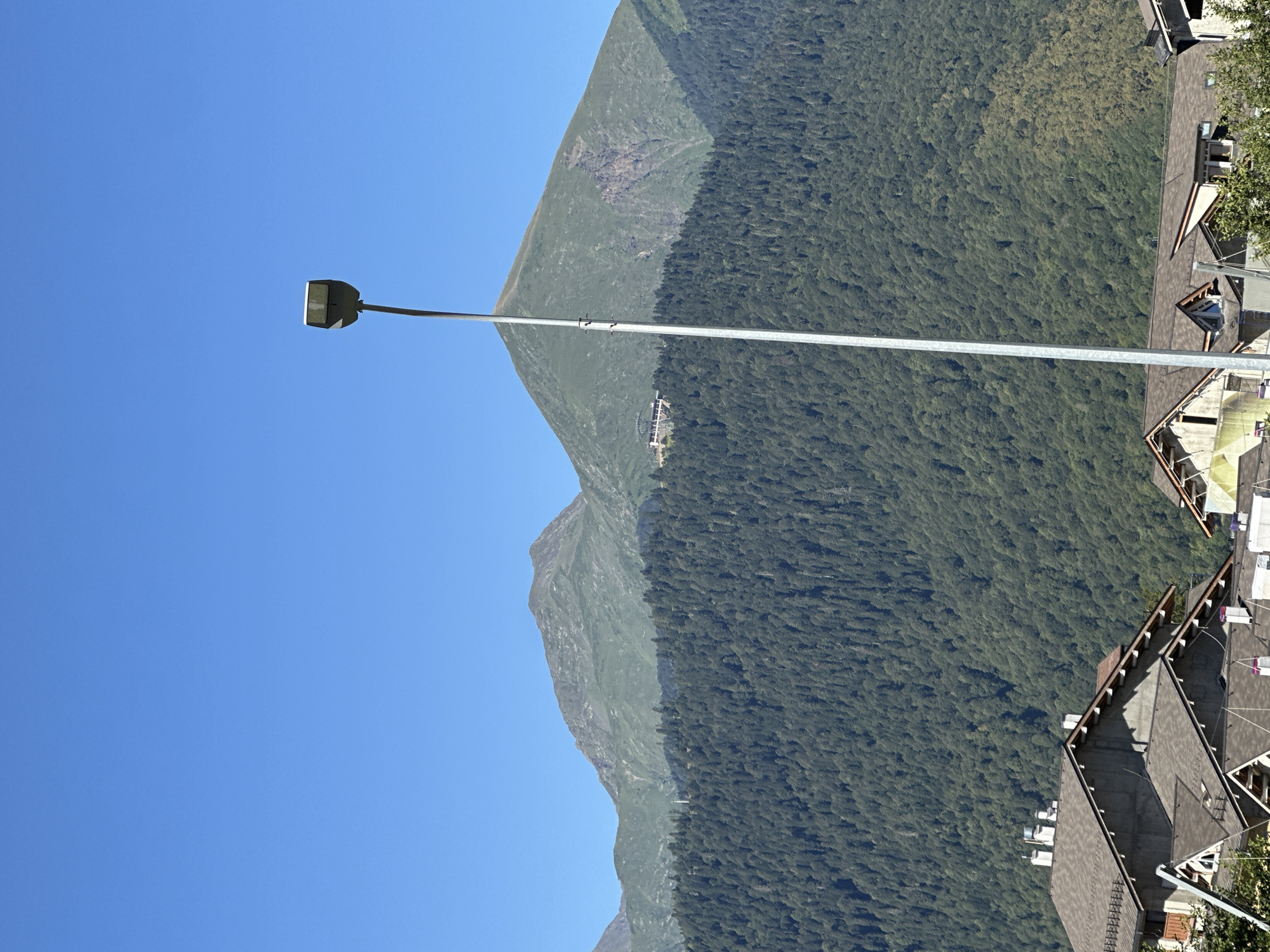 A vertical medium shot captures a clear blue sky above a mountainous landscape dotted with a dense forest and houses. In the foreground, two houses with grey roofs are visible. A tall, grey lamppost stands in the center of the frame, its head tilted to the left. Behind the houses, a vast, dark green forest covers the lower slopes of the mountains. The upper parts of the mountains are a lighter green, and a building with a light-colored roof is visible on one of the peaks. The sky is a uniform, cloudless blue.
