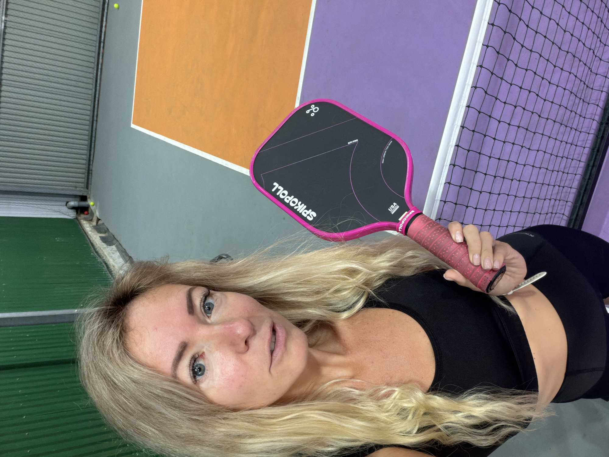 The image shows a woman holding a pickleball paddle, standing on a pickleball court. The court has purple and orange sections, separated by white lines, and a net in the foreground. The background features a green wall and a corrugated metal wall. Several pickleballs are visible on the court. The woman has blonde hair and is wearing a black sports bra and black leggings. She is looking at the camera. The scene is indoors, likely a dedicated pickleball facility. The location in Da Lat, Vietnam is not specified in the image.