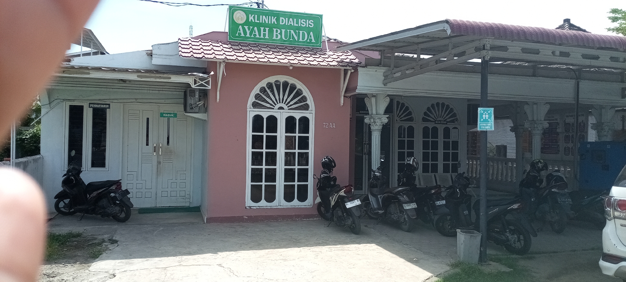 The image depicts the exterior of a medical clinic in Lhokseumawe, Indonesia. The clinic, named "Klinik Dialisis Ayah Bunda" (Dialysis Clinic Father Mother), has a green sign with white lettering above the main entrance. The building consists of three distinct sections: a white section on the left with windows labeled "PENDAFTARAN" (Registration) and a "MASUK" (Enter) sign on the door; a pink section in the center with a large white decorative window labeled "72 AA"; and a larger, gray section on the right with a covered entrance supported by columns. A sign indicating an assembly point is visible near the right entrance. Several motorcycles are parked in front of the clinic, and part of a white vehicle is visible on the right side of the frame. The scene is captured in daylight.