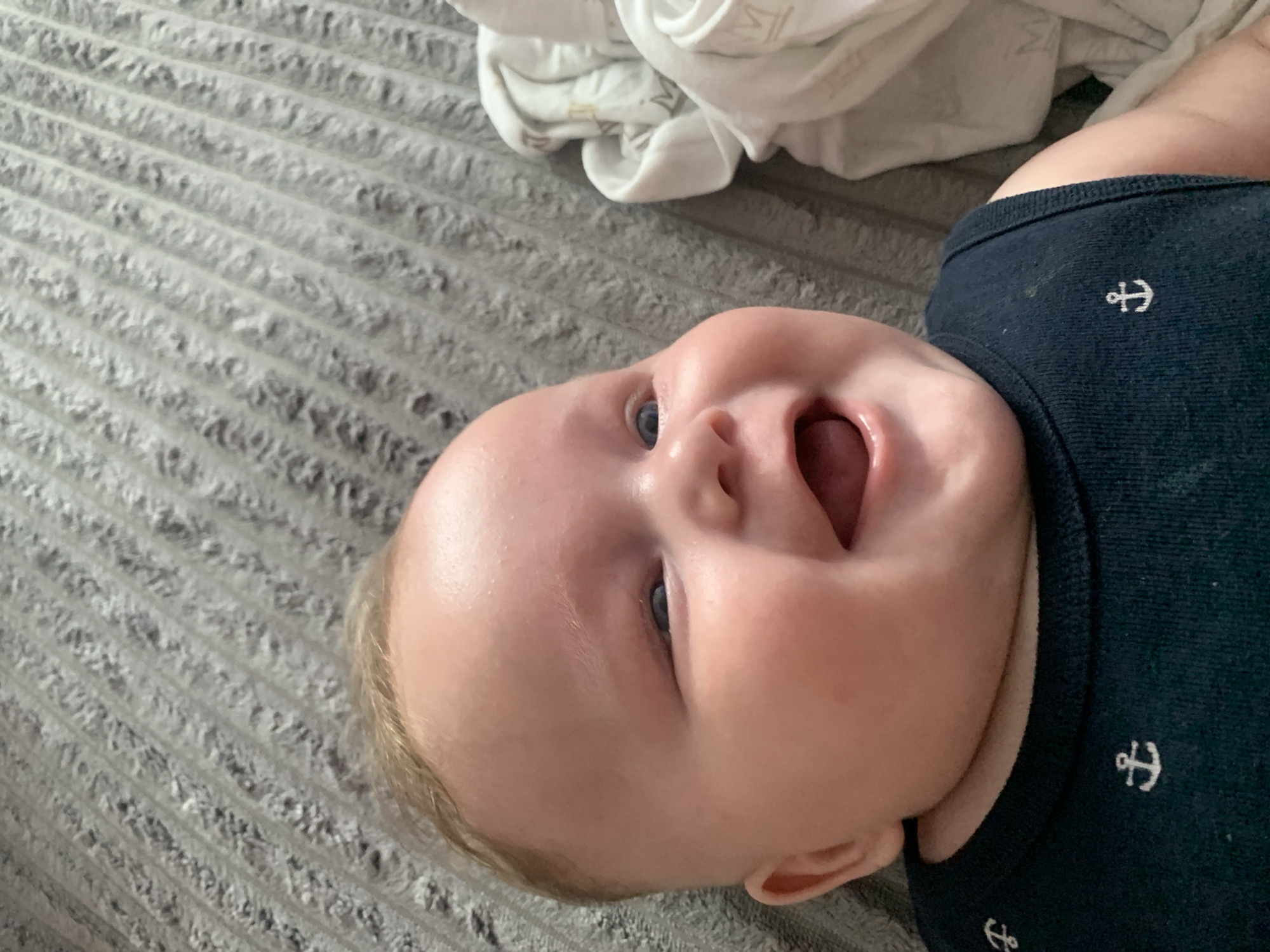 A baby with fair hair and blue eyes is lying on a gray, ribbed surface, possibly a blanket or bed. The baby is smiling widely, with its mouth open and tongue visible. The baby is wearing a dark blue sleeveless shirt adorned with small white anchor motifs. To the right of the baby, a white fabric with a gold pattern, possibly a blanket or swaddle, is visible. The scene appears to be an indoor setting. There is no visible indication of the city of Stavropol, Russia.