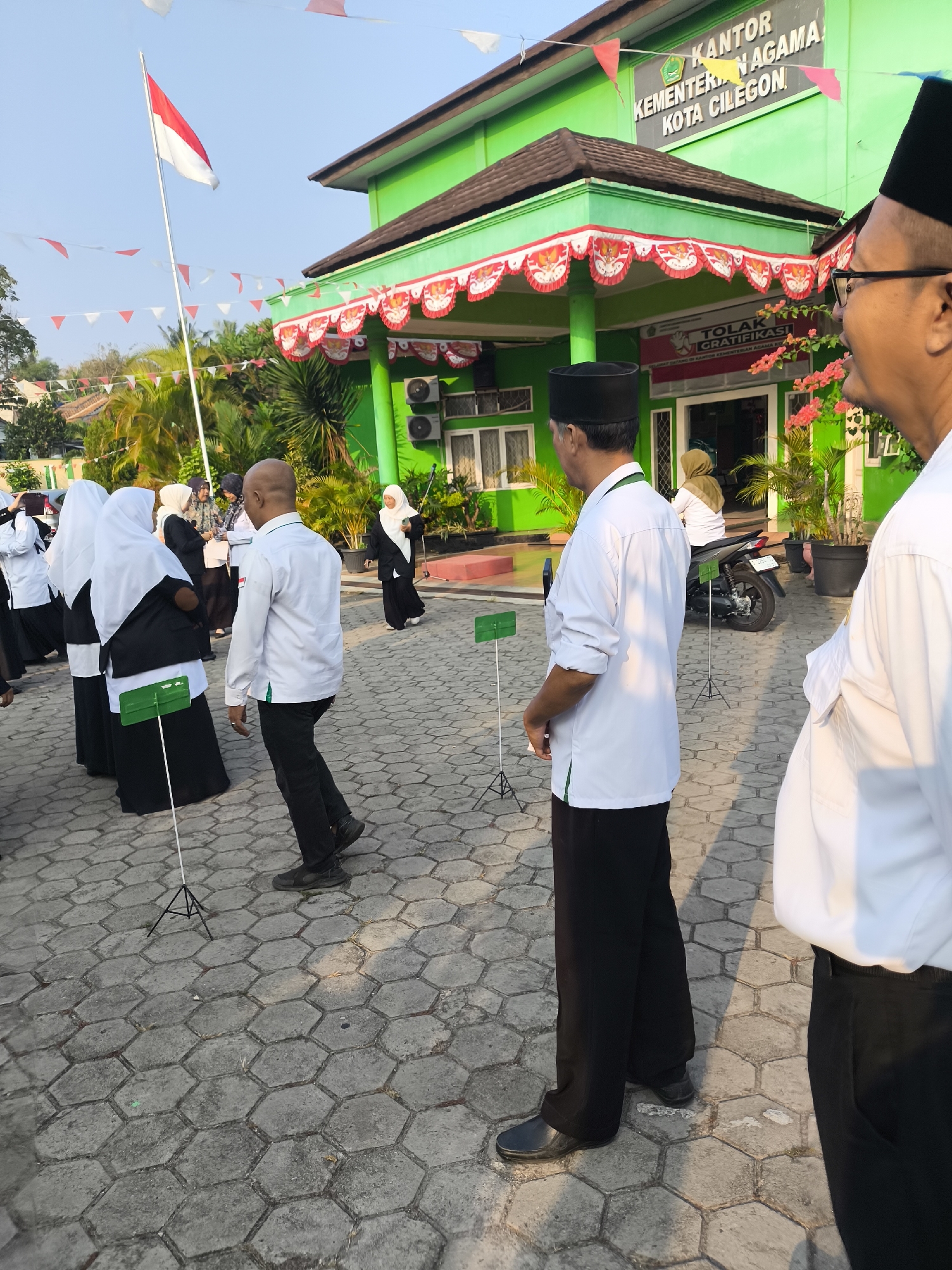 The image depicts the front exterior of the "KANTOR KEMENTERIAN AGAMA KOTA CILEGON" (Ministry of Religious Affairs Office, Cilegon City), located in Cilegon, Indonesia. The building is painted bright green with a brown-tiled roof and features white trim and red and white decorative banners along the roofline. Flags, including an Indonesian flag, and triangular paper flags in red and white are visible, suggesting a national holiday or celebration.
Several individuals are present in the courtyard area in front of the building. In the foreground, two men are partially visible from the back, both wearing black peci (caps) and white long-sleeved shirts with dark trousers. One man in the foreground on the right is looking to the side. Behind them, a group of people, mostly women wearing hijabs and black and white attire, are gathered. Another man in a white shirt and black peci stands facing away from the camera. In the middle ground, a person is seated on a black motorcycle. The ground is paved with hexagonal gray stones. Small black stands with green signs are placed at intervals on the ground. The scene appears to be outdoors during daylight, with sunlight casting shadows.