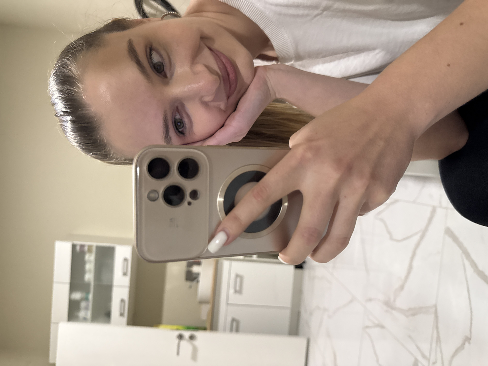 A woman is taking a selfie with an iPhone. The background appears to be a bathroom with white cabinets and a marble floor. The location is unlikely to be determined as being in the city of Budva, Montenegro from this image.