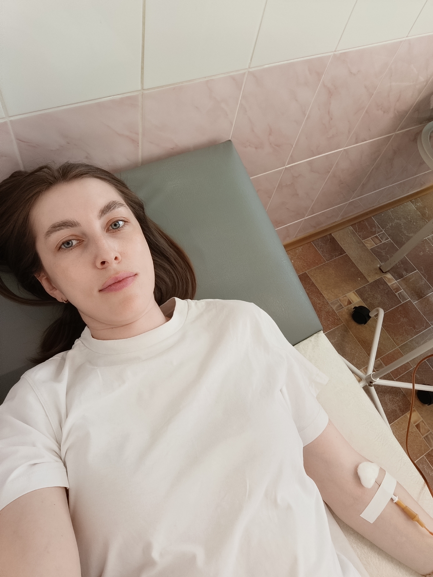 A young adult woman lies on an examination table in a medical facility. She wears a white t-shirt and has an intravenous line inserted in her left arm, secured with a bandage and cotton ball. The room features white and pink tiled walls and a patterned tile floor. Medical equipment, including a stand on wheels with tubing, is visible near the foot of the table. The scene is likely located in Chaikovsky, Russia.
