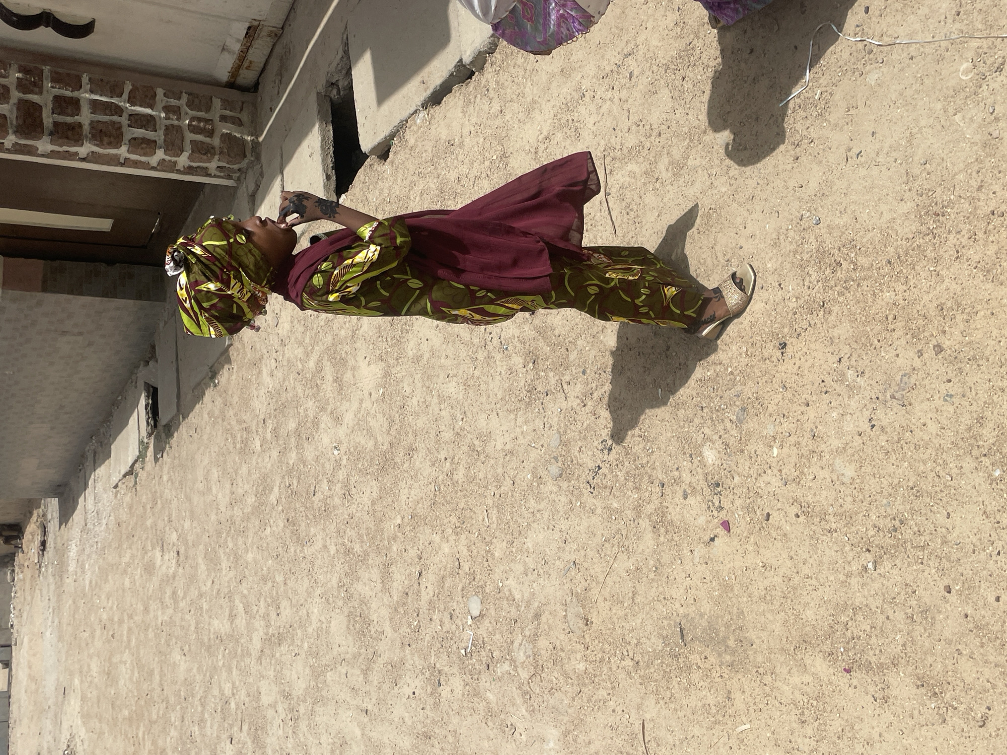 The image depicts a person standing in front of a building on a dirt ground. The person is wearing a patterned green and yellow outfit with a dark red shawl, matching head wrap, and gold shoes. They are near a building constructed with brick and concrete. The location is assumed to be in an urban environment in the city of Unknown, Unknown. The person appears to be putting something in their mouth.