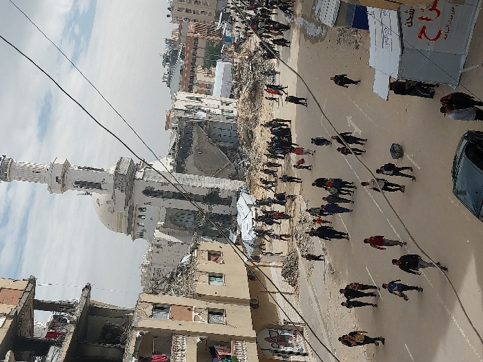 The image depicts a street scene in Khan Yunis, Palestinian Territory, characterized by significant structural damage and a high volume of pedestrian traffic under an overcast sky. In the mid-ground, a prominent mosque, likely the Great Mosque of Khan Yunis, stands partially destroyed, with its white minaret and dome largely intact but significant portions of its lower structure collapsed into rubble. To the left of the mosque, a multi-story residential building shows extensive damage, with exposed rebar, shattered windows, and laundry hanging from a damaged balcony.
The paved road in the foreground and mid-ground is densely populated with dozens of individuals, including adults and children, walking in various directions. Many appear to be carrying bags, backpacks, or small pieces of luggage, suggesting displacement or movement of personal belongings. Piles of debris and rubble are visible along the sides of the road and in the background, where more damaged buildings can be seen. Several makeshift tents or shelters, some white, others with Arabic writing, are set up along the right side of the road. Overhead, multiple black power or utility lines traverse the scene. A dark-colored sedan is parked in the lower right foreground. The overall environment conveys a scene of post-conflict or disaster.