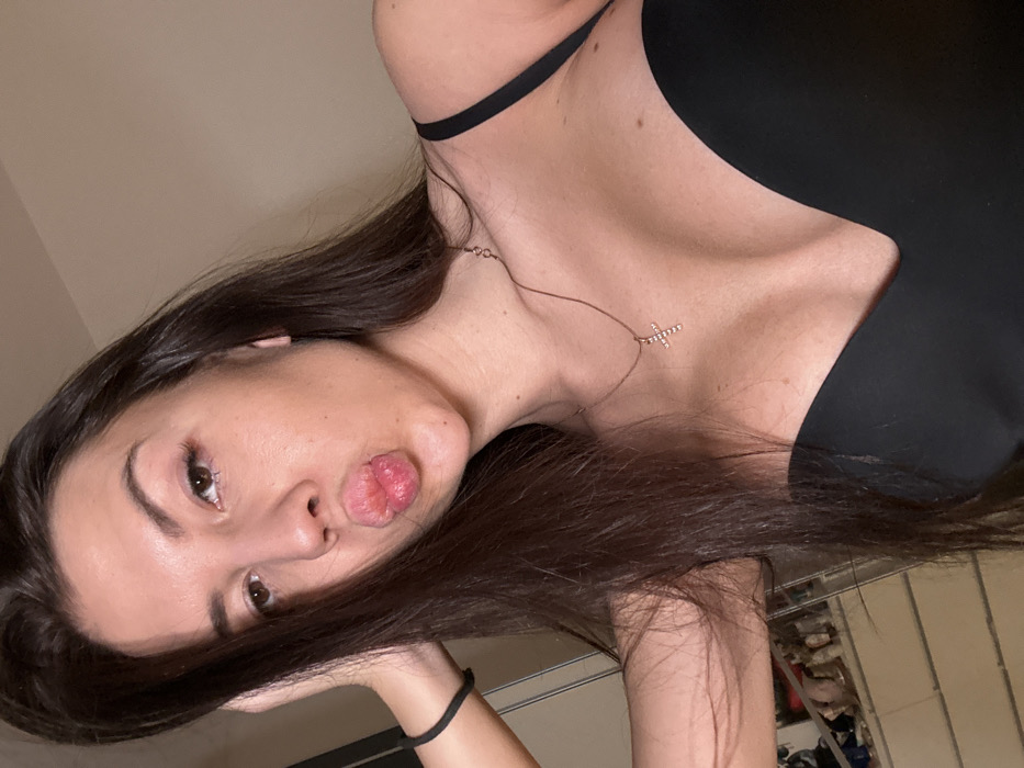 A woman with long, dark brown hair poses for a selfie. She is wearing a black tank top and a delicate necklace with a cross pendant. Her face is tilted to the side, and she is puckering her lips. Her left arm is slightly visible, showing a black elastic band on her wrist. The background is a light beige wall, with a glimpse of a rack of shoes partially visible behind her head.