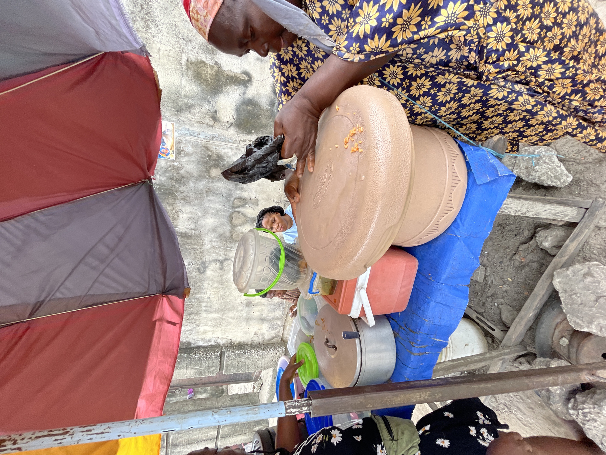 In Ado, Nigeria, a woman wearing a vibrant yellow and navy blue floral patterned dress and a red headscarf is captured at an outdoor food stall. She is holding a black plastic bag in her right hand and is in the process of placing food into a large, tan-colored, plastic container that sits on a blue tarp covering a table. Another woman, dressed in a black shirt with a white floral pattern and green cargo pants, stands to the left, facing away from the camera, her arm extended towards the food containers on the table. A younger person, a girl wearing a light blue t-shirt, is visible behind the food containers, looking towards the woman handling the food. The stall is set up outdoors, with a red and gray umbrella providing shade. The background consists of a rough, concrete wall. On the table are various food containers, including metal pots, plastic tubs, and a red cooler. The ground beneath the stall is dirt and rocks.