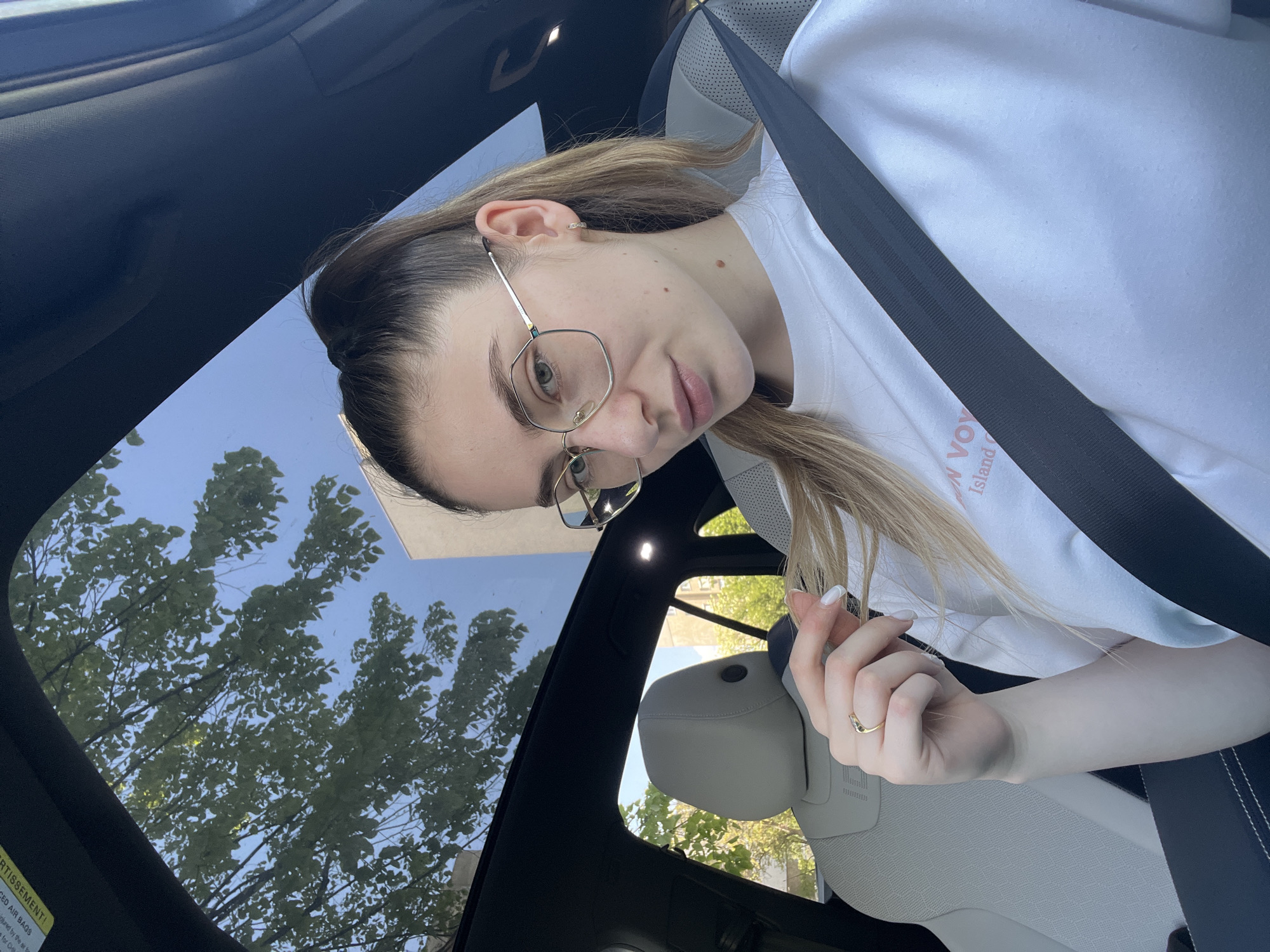 A medium-shot photograph shows a woman with brown hair in a high ponytail, wearing glasses and a white t-shirt, sitting in the driver's seat of a car and wearing a seatbelt. The car's sunroof offers a view of trees and a blue sky. The location is Unknown, Unknown.