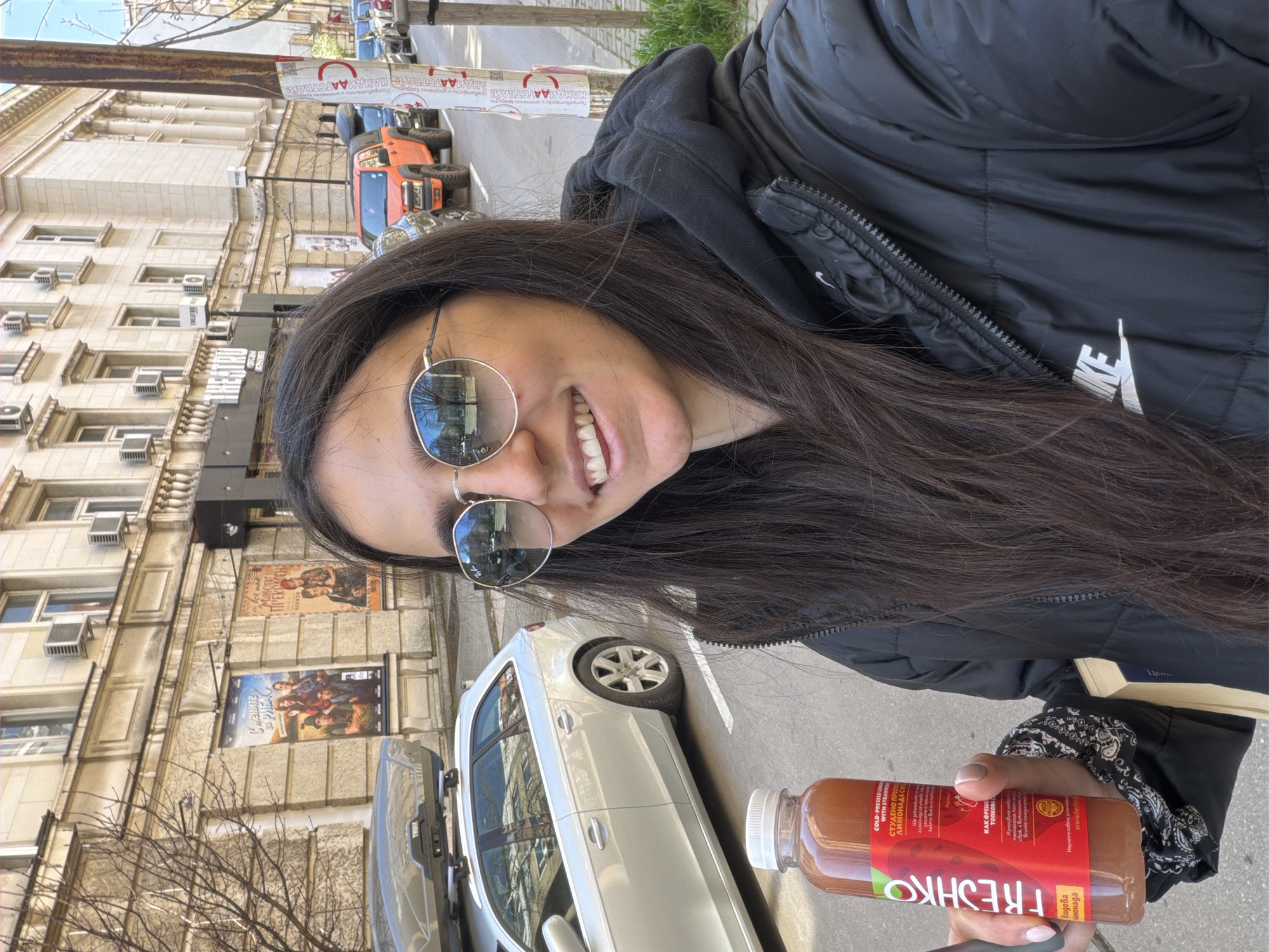Here is a detailed description of the image:
**Overall Scene:** The image appears to be a selfie taken outdoors, likely in an urban setting in Sofia, Bulgaria. The orientation of the photo suggests it was rotated to appear upright.
**People:** The main subject is a young woman. She has long, dark hair and is wearing reflective sunglasses and a black Nike jacket with a hood. She's smiling.
**Objects:**
* A bottle of a "Freshko" drink is held in the woman's hand.
* A white car is parked behind the woman.
* Buildings are visible in the background, including one with multiple floors and what appear to be air conditioning units in the windows, and movie posters
* Various other cars are parked on the street in the background.
* A pole with advertisements/signage is also present.
**Environment:** The setting is a street in Sofia. The presence of parked cars, buildings, and advertisements suggests a relatively busy urban area. The bare trees hint it might be late winter or early spring.
**Details Indicating Sofia Location:**
* The writing on the advertisements/signage on the pole appears to be in Cyrillic, the script used for Bulgarian.
* The product name "Freshko" is common in Bulgaria.
**Actions/Interactions:** The woman is taking a selfie while holding the drink.