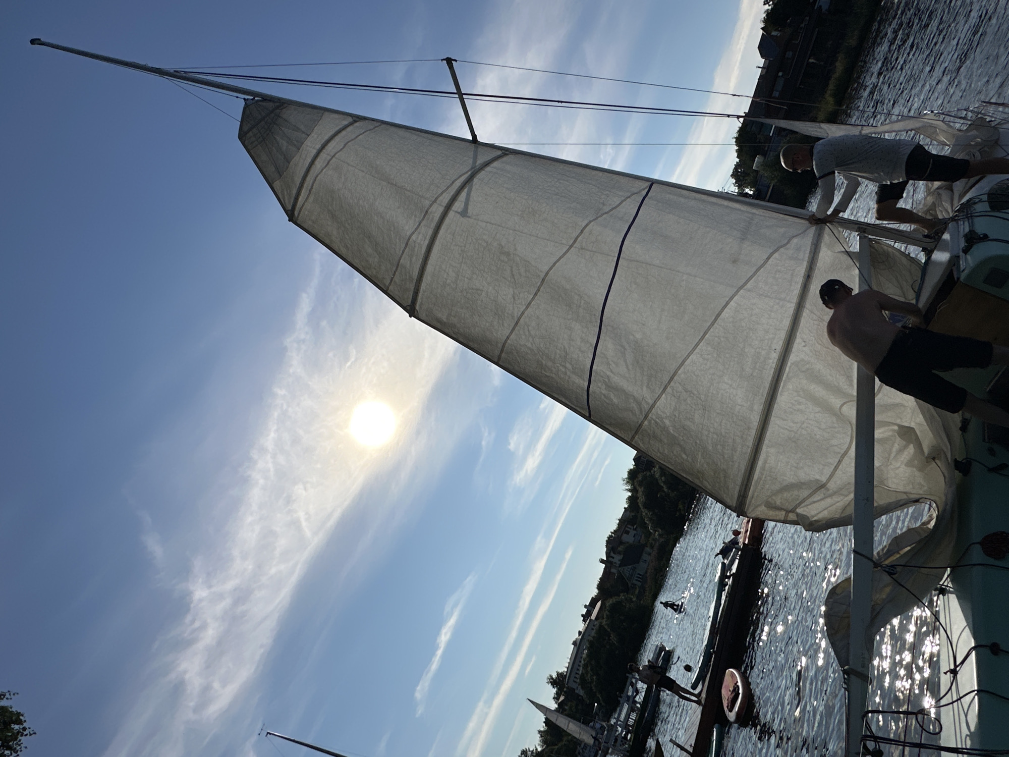 In a low-angle shot at Туристический Пансионат «Клязьминское водохранилище», Russia, two individuals are working on a sailboat on a body of water. The sun, partially obscured by thin clouds, shines brightly in a clear blue sky. A large, white sail, partially unfurled, dominates the right side of the frame, extending upwards towards the sky. The sail has dark blue lines running horizontally across it.
One person, appearing to be a male, is shirtless and wearing dark shorts, leaning over the boat's edge to adjust something. The other person, also seemingly male, is wearing a light-colored t-shirt and dark shorts, actively pulling a rope attached to the sail. The boat itself appears to be a small sailboat, with visible rigging and parts of its deck.
The water is choppy and reflects the sunlight, creating a shimmering effect. In the background, a wooden dock with several paddleboards is visible, with another person standing on it. Further in the distance, trees and houses line the shore, suggesting a developed area along the water. The overall scene suggests an activity related to preparing a sailboat for sailing on a bright, sunny day.