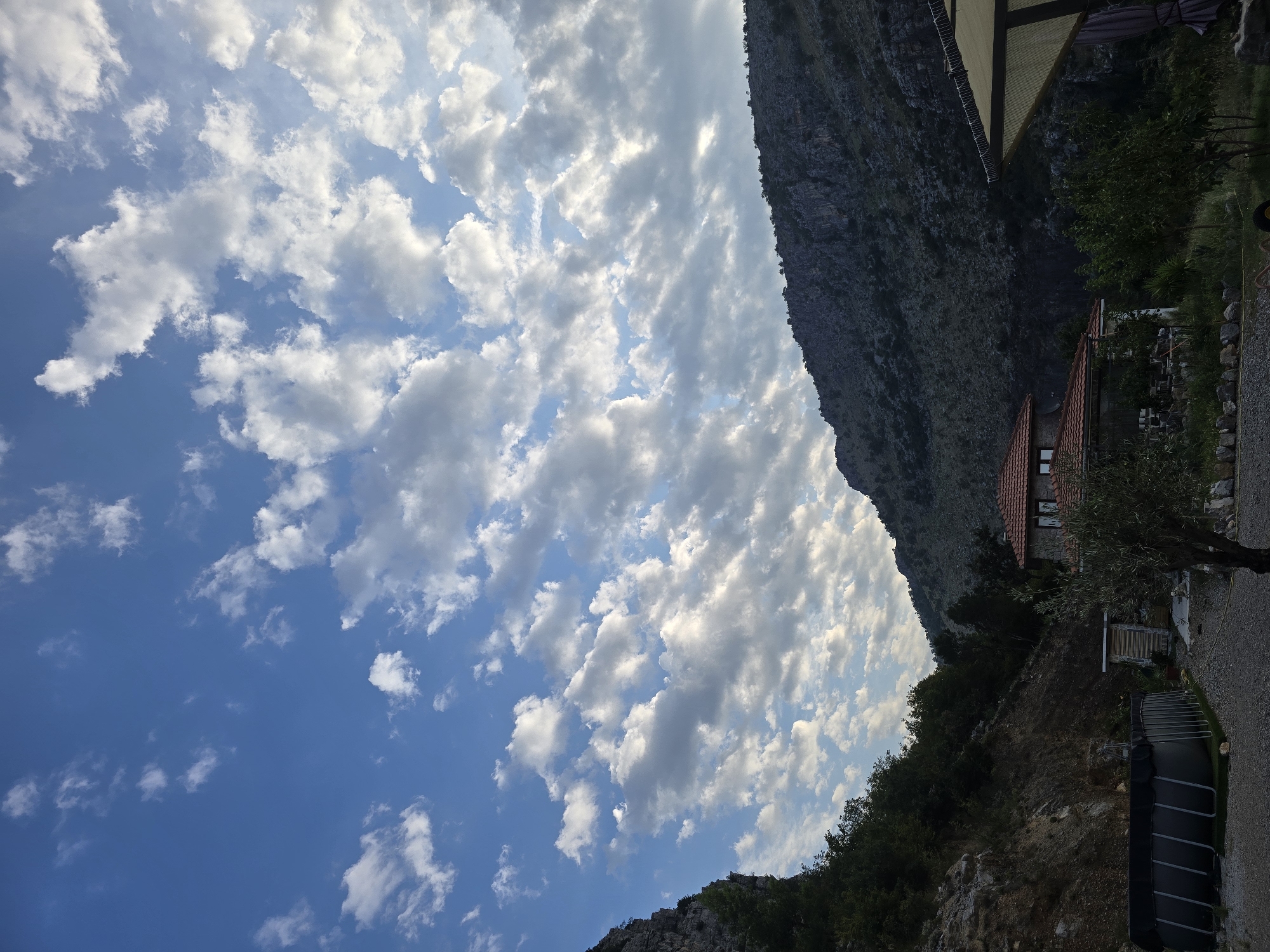 The image depicts a rural landscape with a dwelling in Montenegro, near Podgorica. The scene is dominated by a bright blue sky filled with scattered white clouds. In the foreground, there is a dark, rectangular swimming pool with metal supports, situated on a gravel surface. Adjacent to the pool, a small, wooden shed with vertical siding is visible. Further into the property, a house with a red-tiled roof and stone walls is partially obscured by foliage. A young olive tree stands in front of the house. The background features a steep, rocky mountain slope covered in sparse vegetation. To the right, a portion of an outdoor canopy with a beige fabric roof is visible. The overall impression is a serene, daytime setting in a mountainous region.