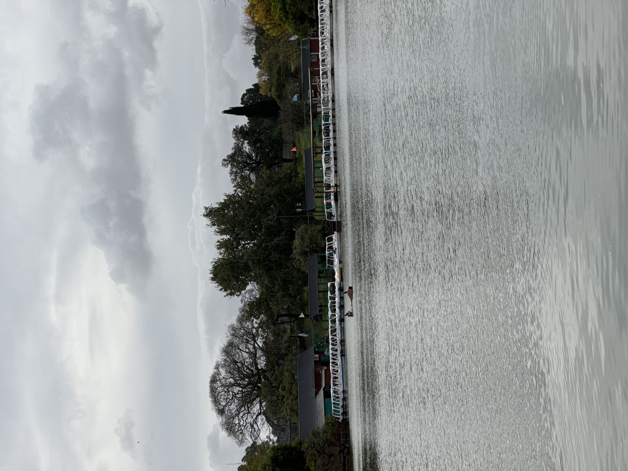 The image captures a view of a lake in Buenos Aires, Argentina on a cloudy day. Along the edge of the lake, there's a series of connected docks or piers, each marked with white railings. Vegetation, including trees and shrubbery, lines the shore. A building with a red wall and a dark roof is visible further back. The lake's surface is rippled, suggesting a slight breeze. The sky is overcast with varying shades of grey clouds.